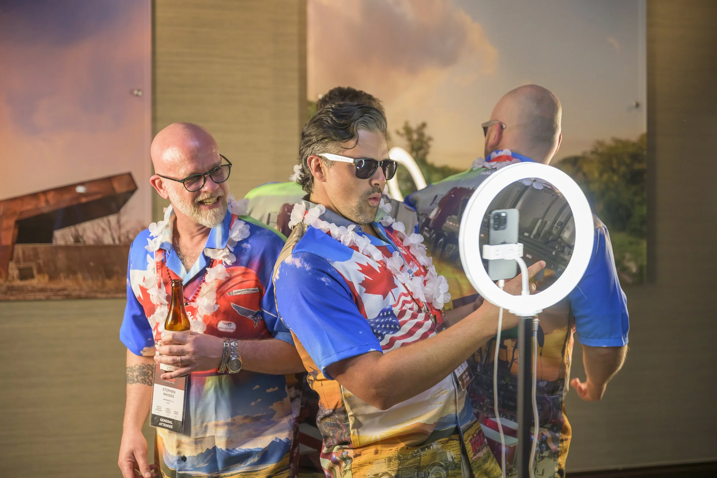 Three men at a party taking a selfie with a ring light. They are wearing colorful, patriotic shirts and leis, with one holding a beer. The background features a sunset scene.