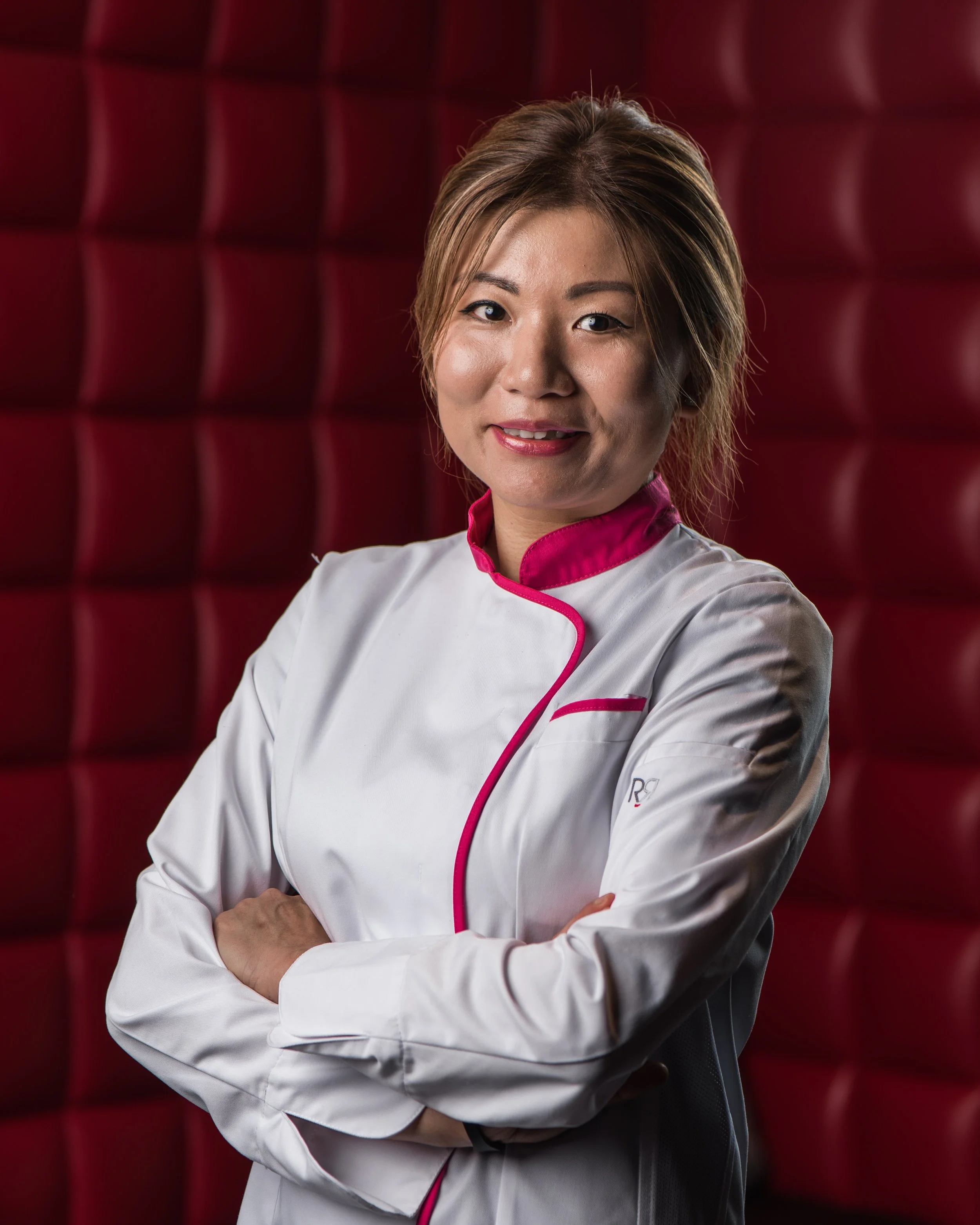 A woman in a white uniform with pink accents, standing with arms crossed in front of a red padded wall, smiling confidently.
