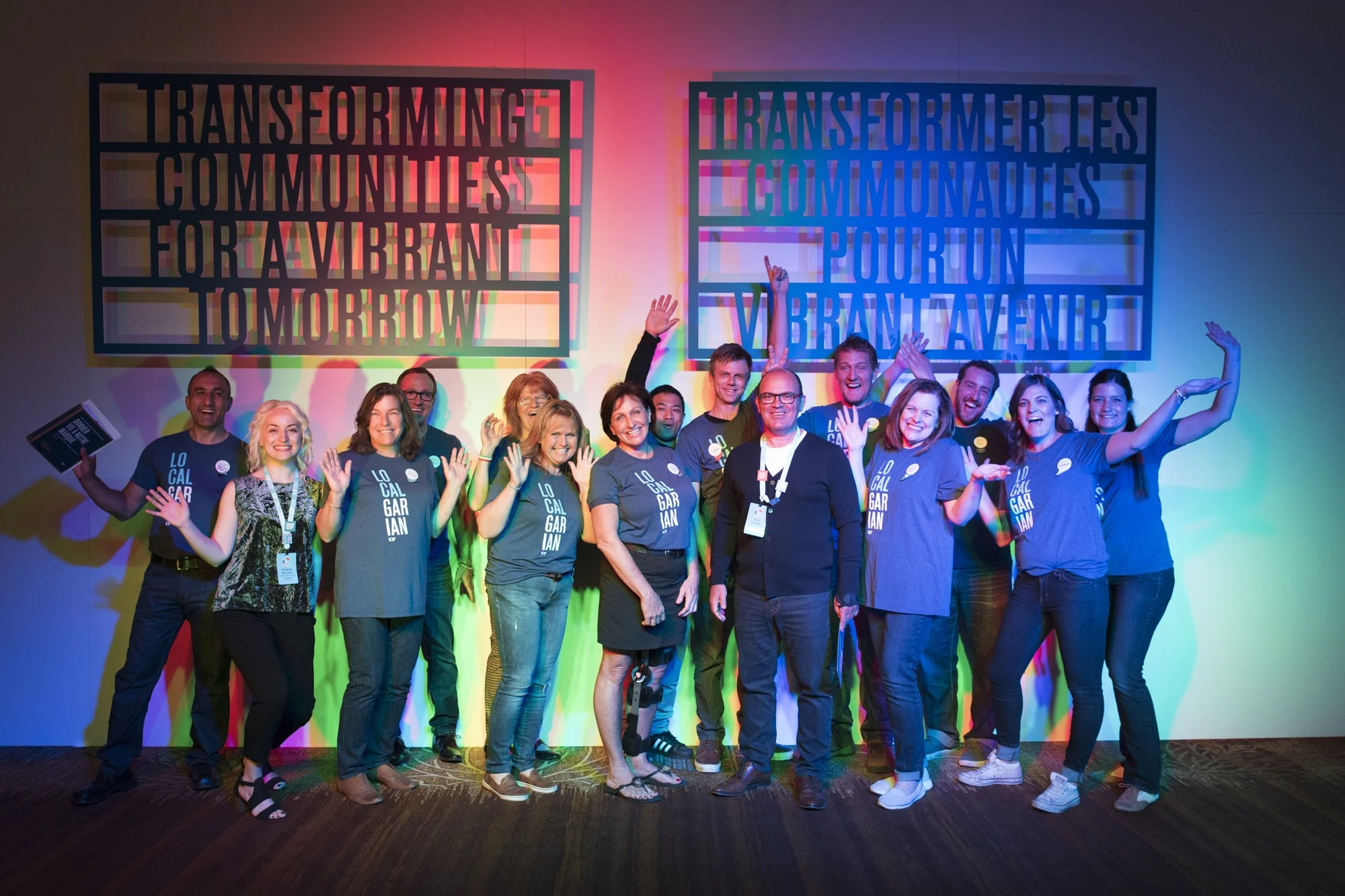 Group of diverse people smiling and raising hands at a conference, with colorful lighting and two large signs in the background with motivational phrases in French and English.