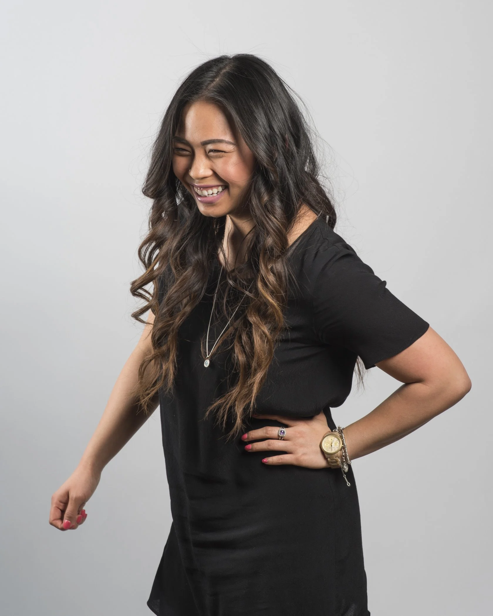 A woman with long, wavy dark hair laughing or smiling, wearing a black dress, a watch, a ring, and a necklace, standing against a plain light gray background.