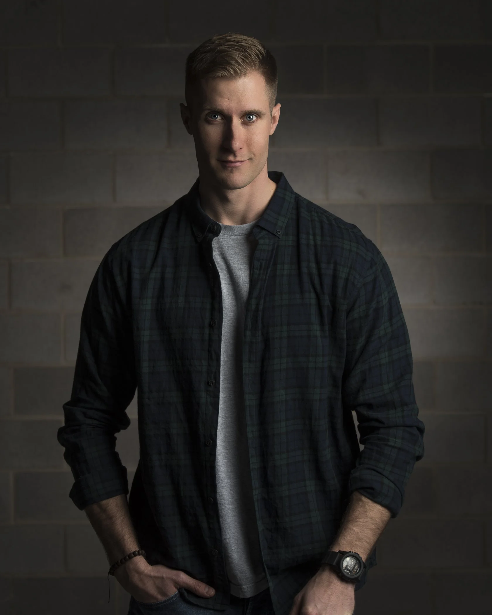 Young man with blonde hair, wearing a dark plaid shirt over a gray t-shirt, standing in front of a dark brick wall, looking at the camera.