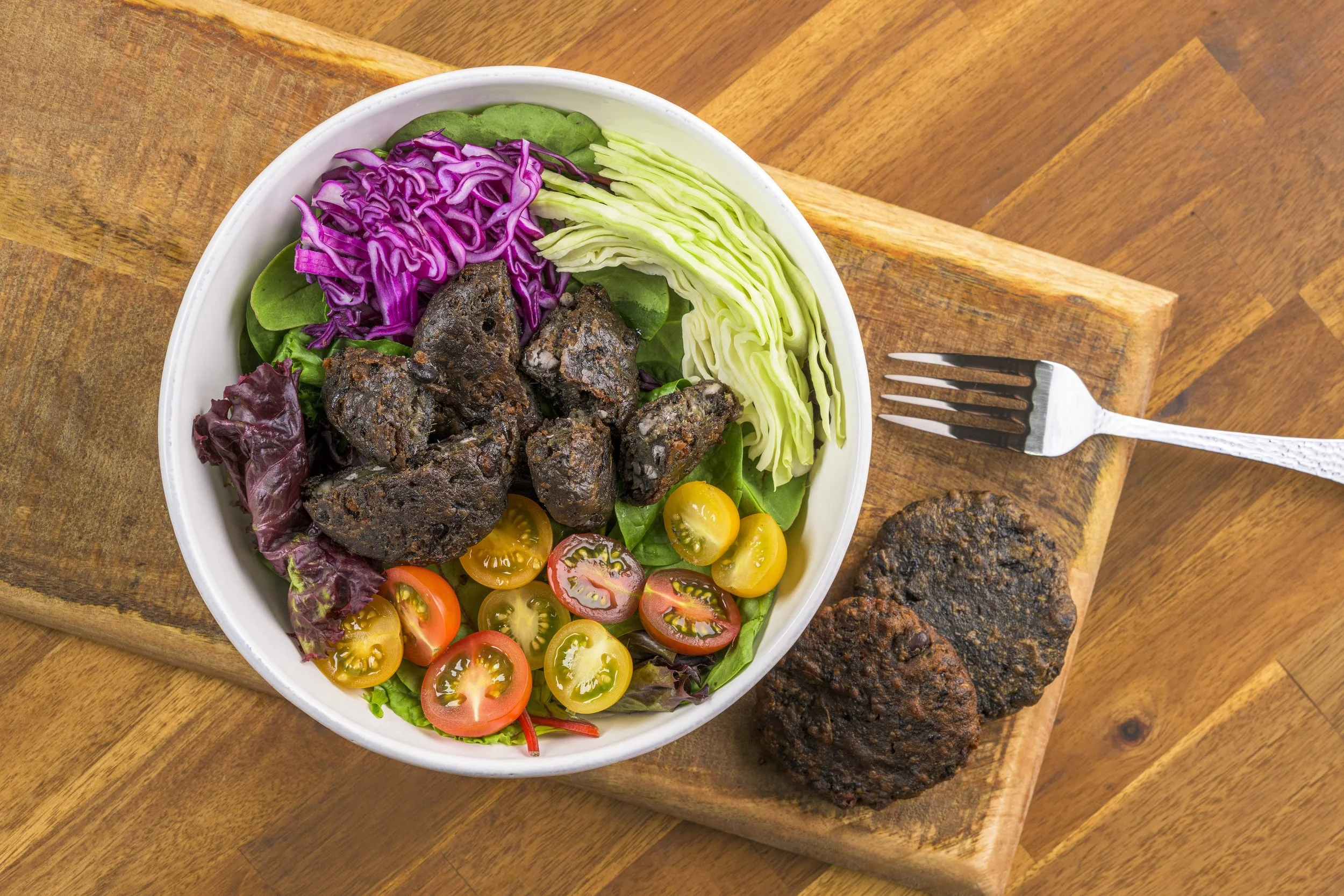 A bowl of salad with purple and green cabbage, cherry tomatoes, and greens, placed on a wooden cutting board, with two veggie burgers next to it.