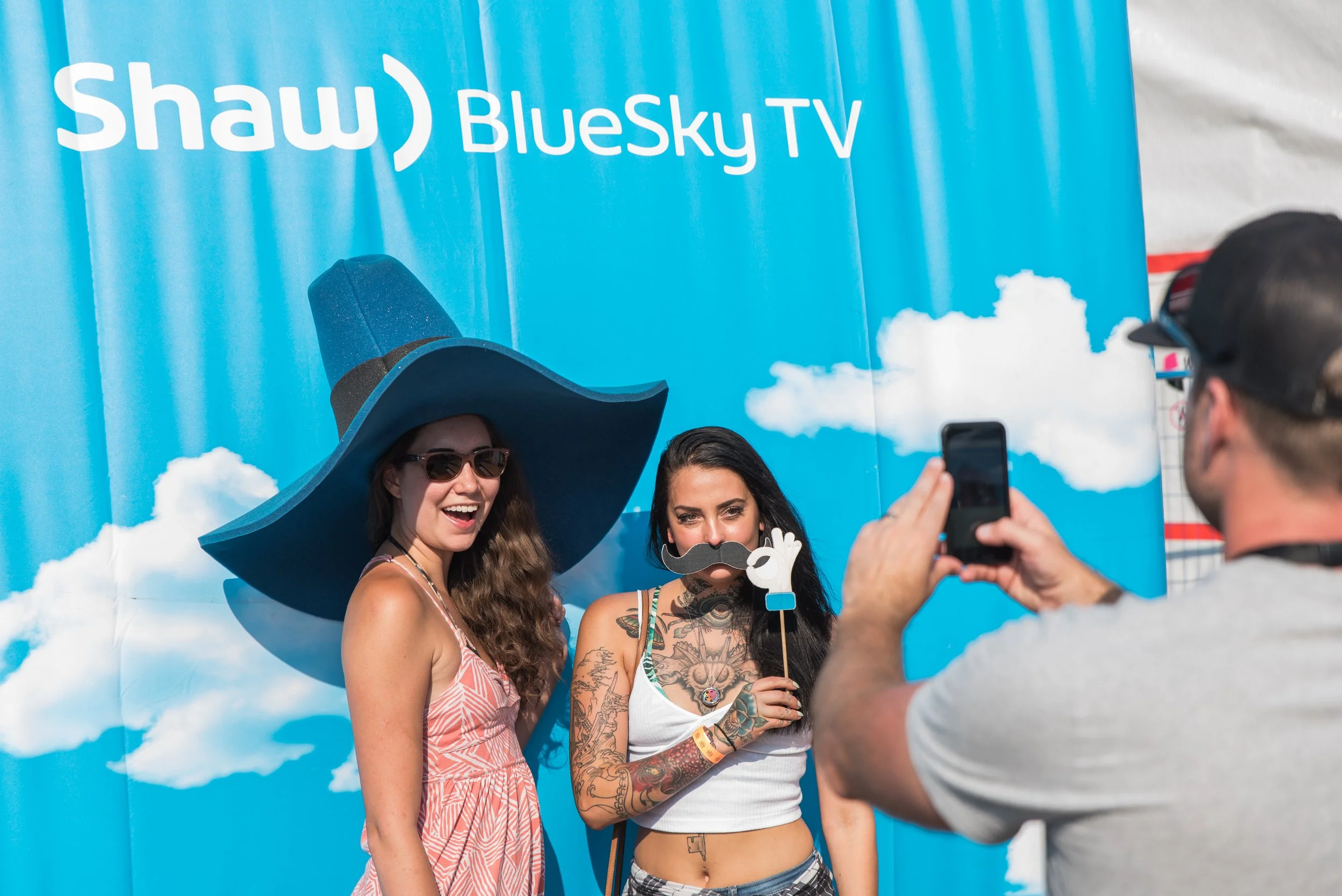 Two women are posing for a photo in front of a blue backdrop with the words 'Shaw BlueSky TV'. One woman is wearing a large blue witch hat and sunglasses, smiling. The other woman has long black hair, tattoos, and is holding a paper mustache and hand