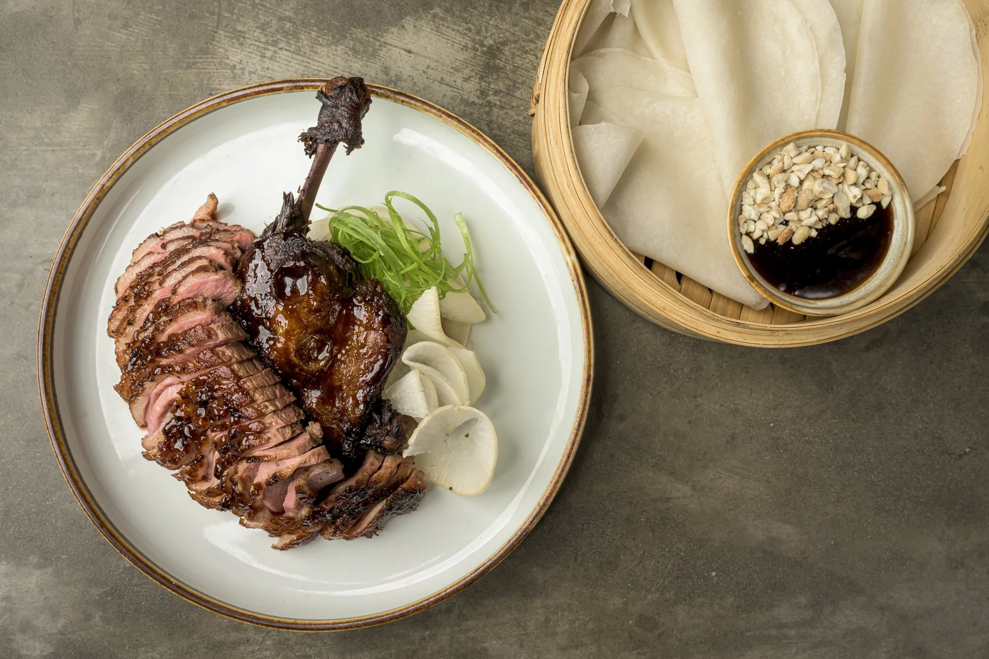 Sliced roasted duck with green onions, white onions, and sauce on a white plate, with steamed rice paper and sauce with chopped peanuts in a bamboo basket.