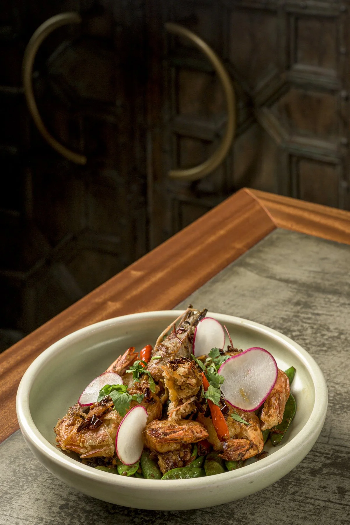Plate of cooked shrimp garnished with sliced radishes, red chili peppers, and herbs on a grey table, with a wooden edge and dark background.