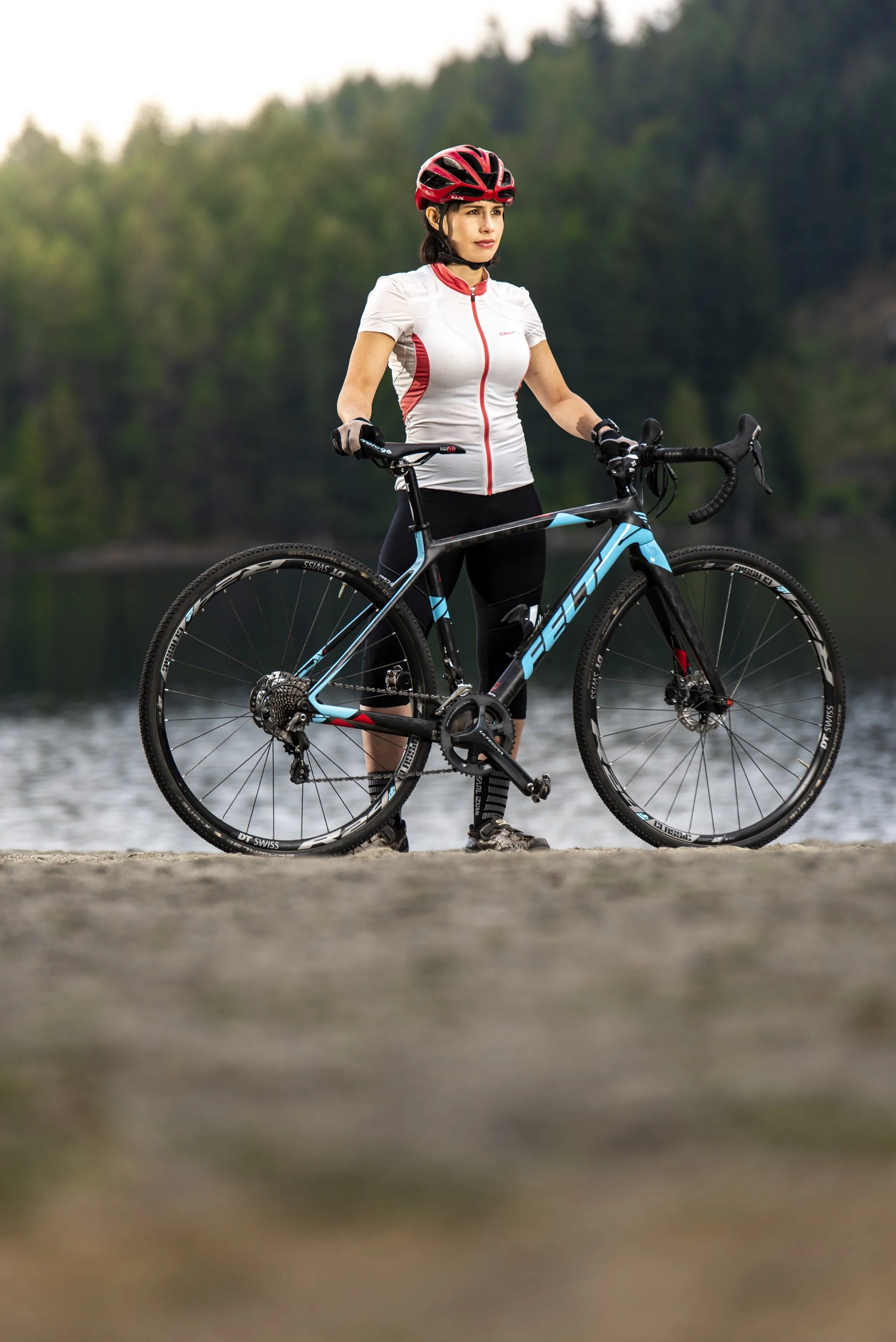 A woman in cycling gear with a red helmet stands outdoors holding a black and blue road bicycle near a body of water, with a forested background.