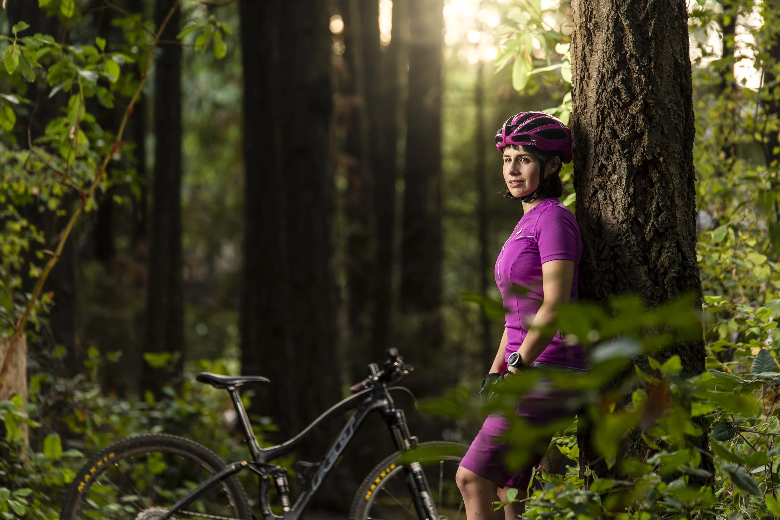 A woman wearing a purple athletic outfit and helmet rests against a tree with a mountain bike in a dense forest during daylight.