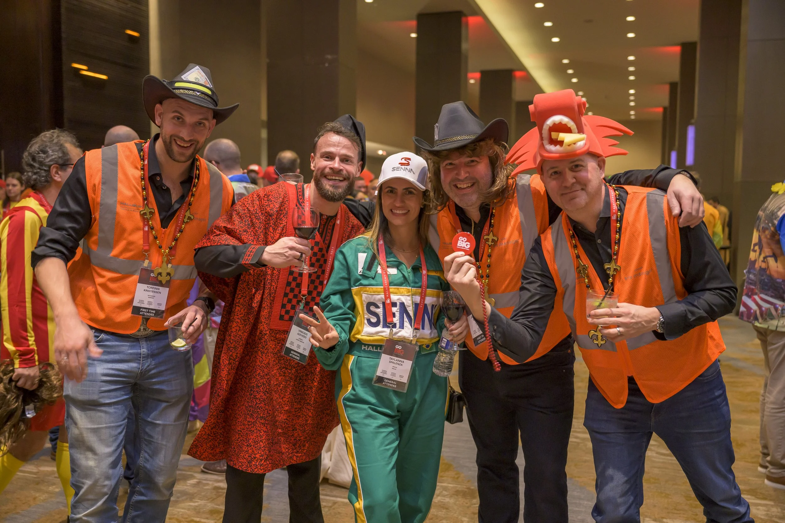 Group of people at a celebration wearing costumes, one person is dressed as a race car driver, others in colorful outfits, some with cowboy hats, one with a dragon hat, all smiling and holding drinks in a large indoor hall.