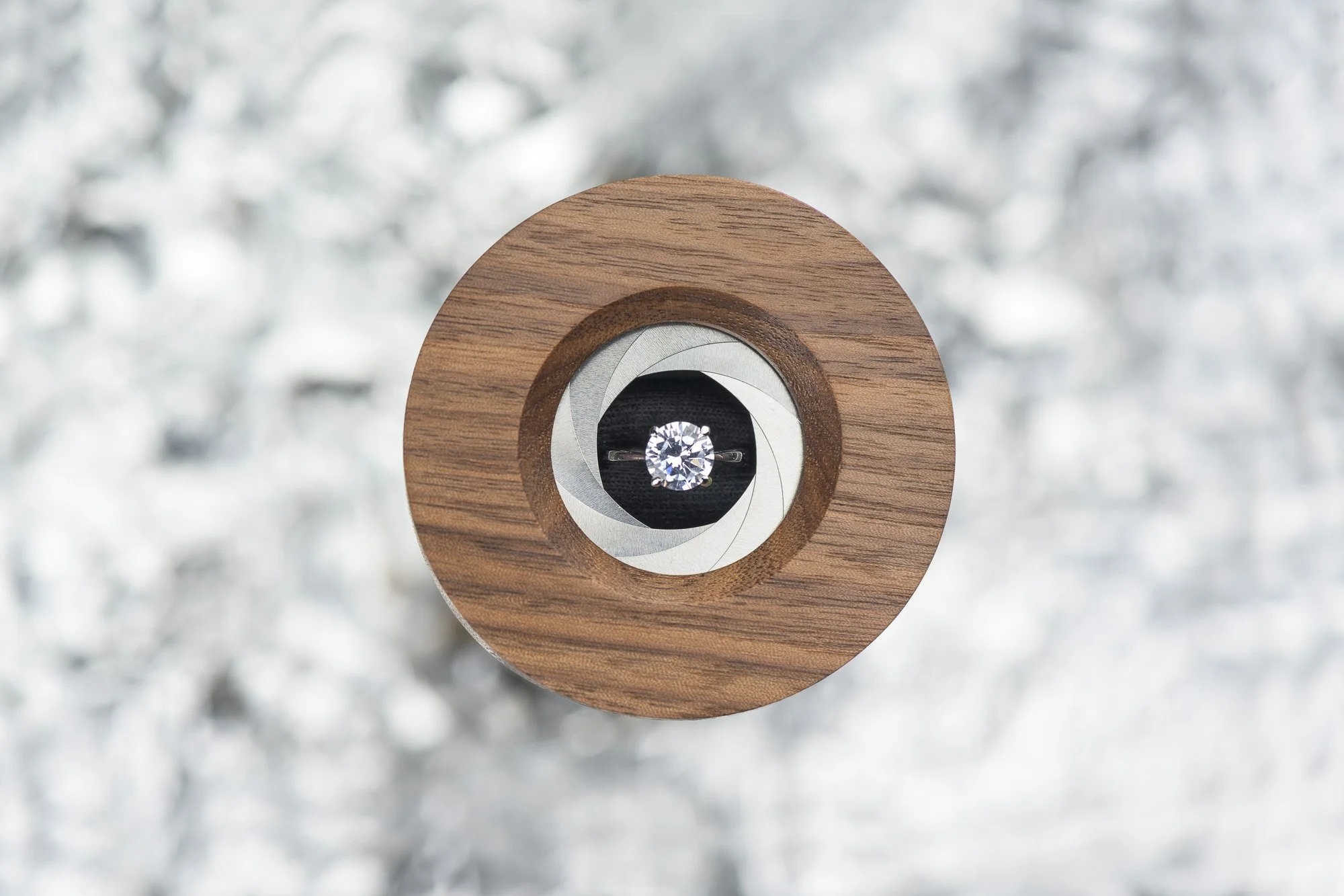 A close-up of a diamond ring set in a circular indentation of a wooden object, against a blurred metallic or shiny silver background.