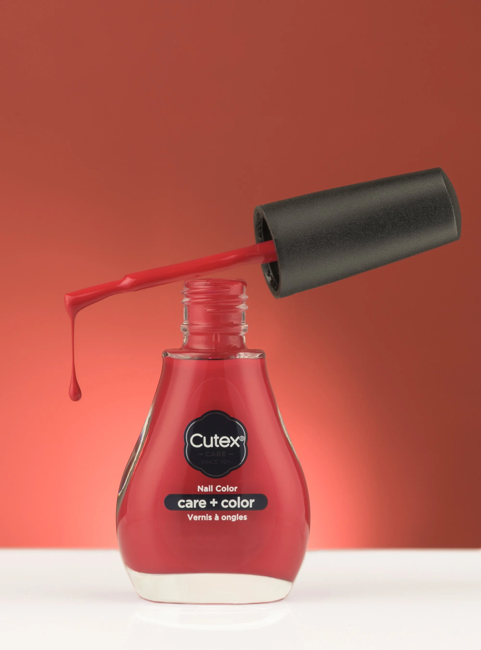 A bottle of Cutex nail polish with a black cap and red polish, with the brush leaning against the bottle, dripping red nail polish.