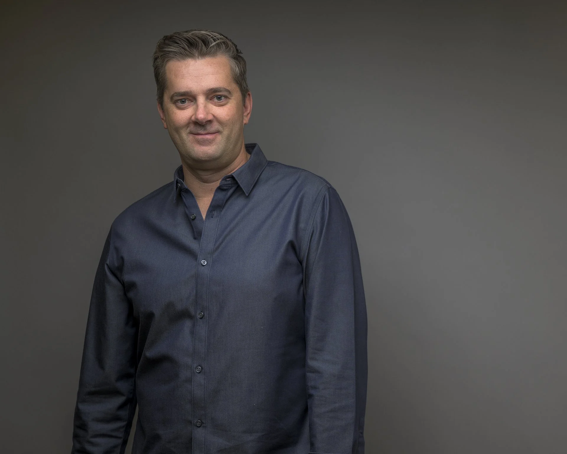 Portrait of a man with light brown hair in a dark button-up shirt, standing against a gradient gray background