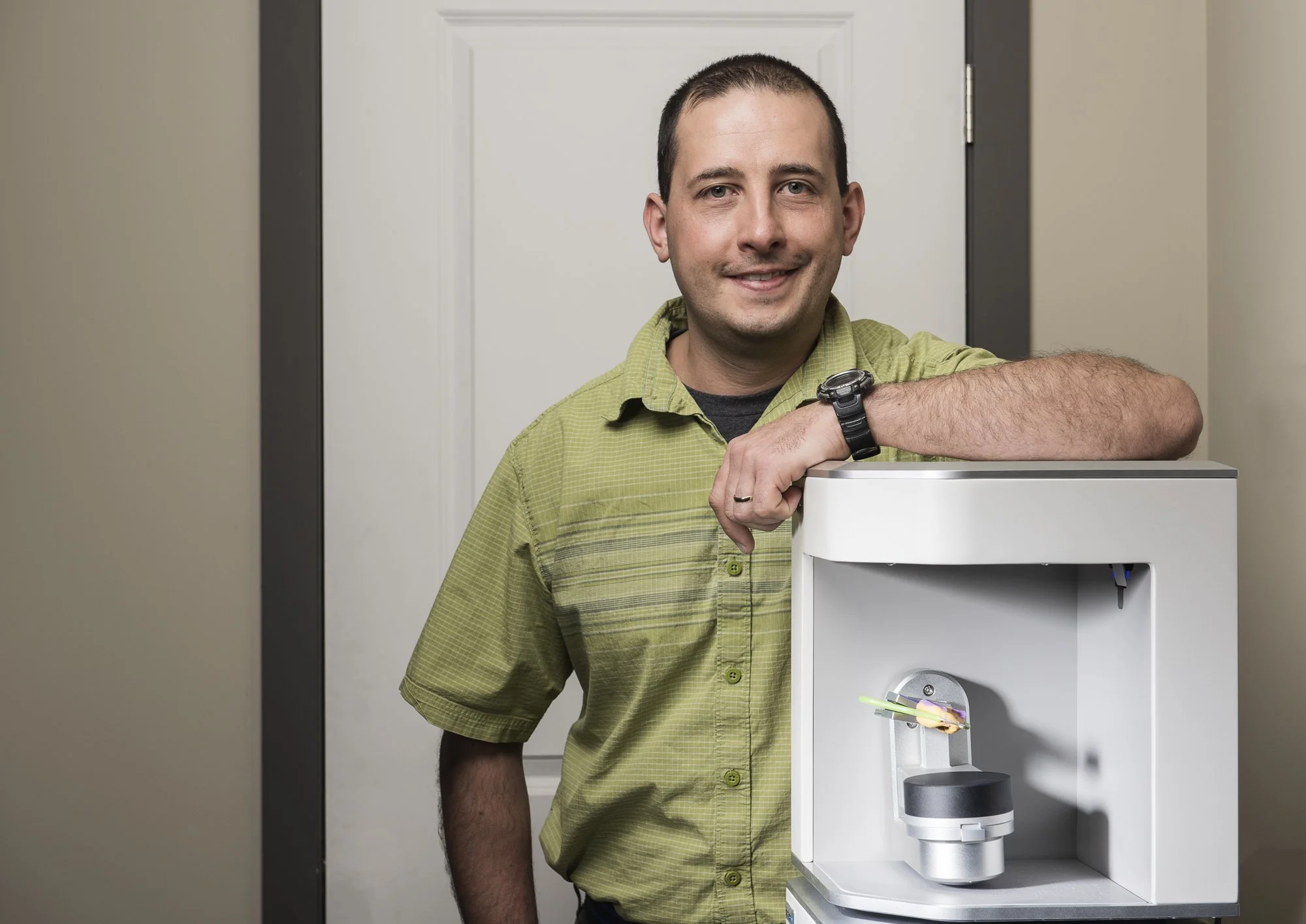 A man in a green shirt smiling and posing with a 3D printer in a room.