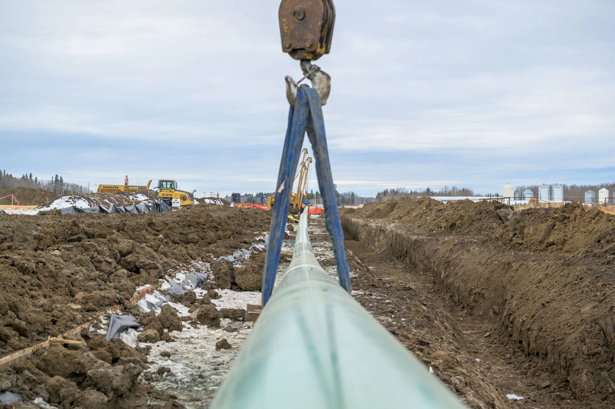 Construction site with a large pipe being installed, heavy machinery, and soil mounds under a cloudy sky.