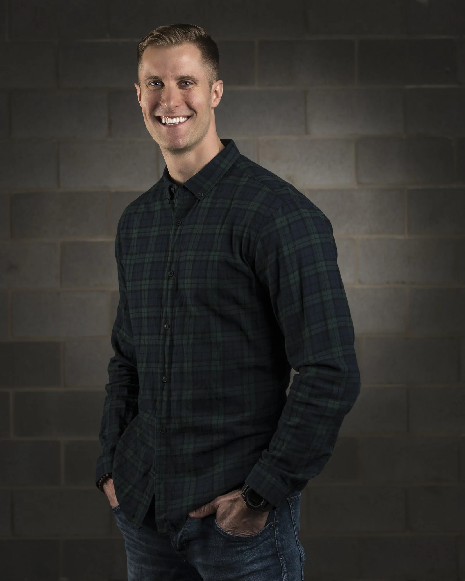 A young man with short blonde hair, smiling, wearing a dark plaid shirt and jeans, with hands in pockets, standing in front of a gray brick wall.