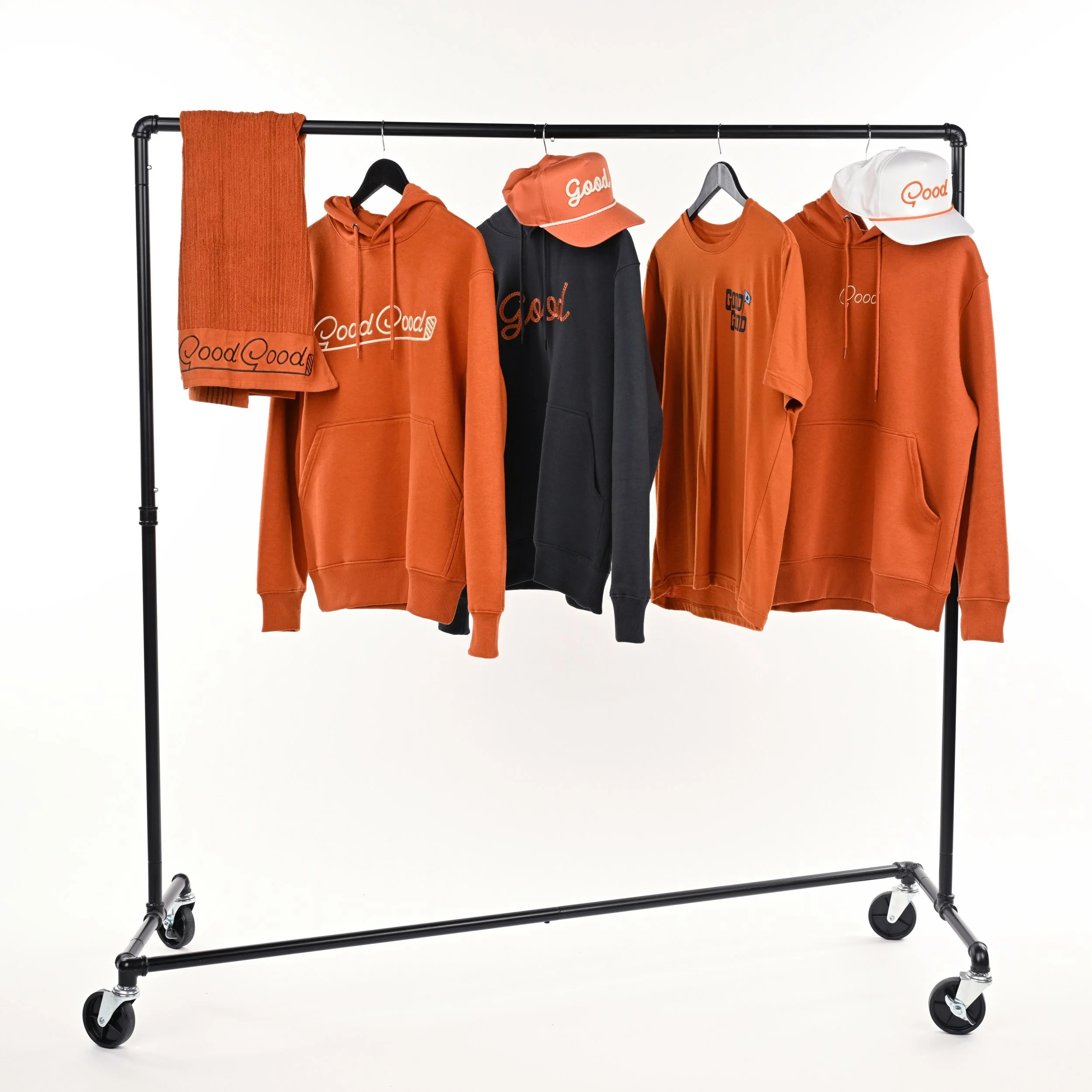 Clothing rack with orange and black hoodies, t-shirt, cap, and scarf, all with 'Good' logo.