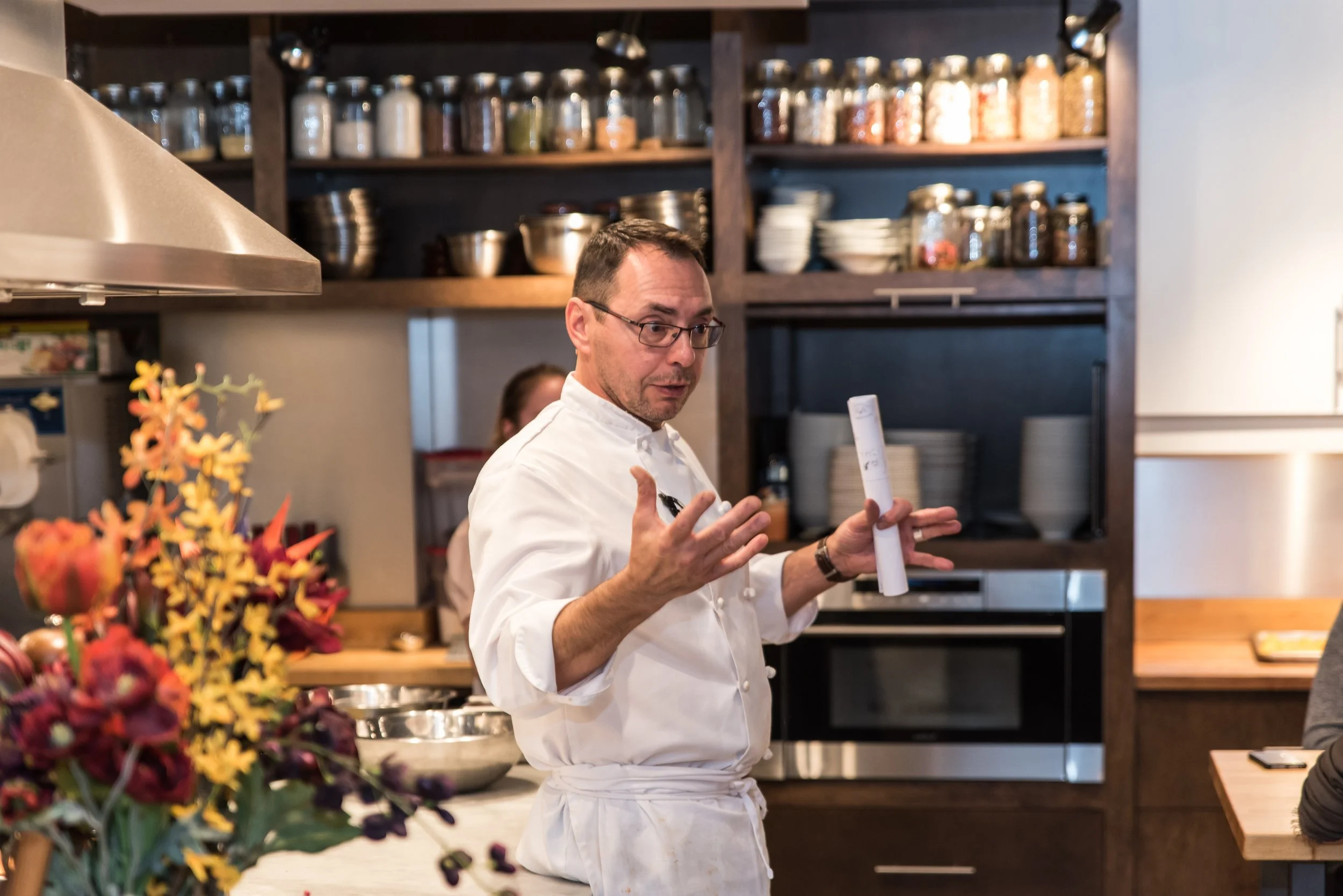 Chef in a kitchen holding a rolled-up recipe, gesturing with his hands while speaking, with a bouquet of colorful flowers in the foreground and shelves with jars and bowls in the background.