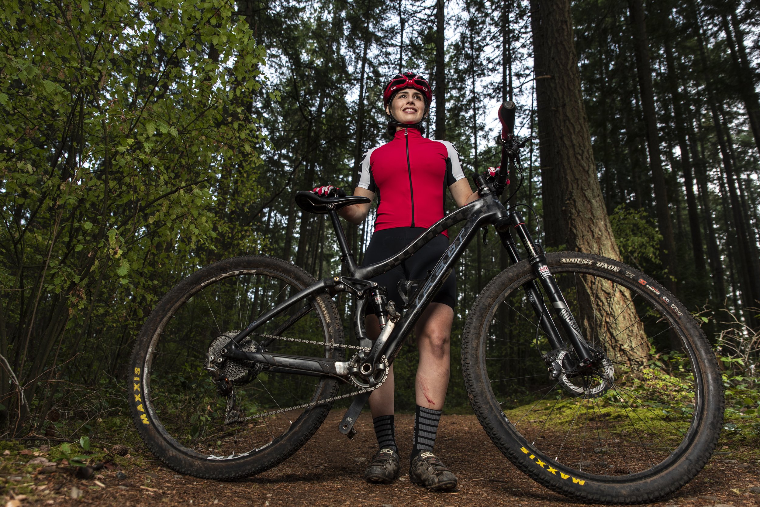 A woman wearing a red cycling jersey, black shorts, and a helmet stands in a forest holding a black mountain bike with yellow Maxxis tires. She appears to have minor scratches on her leg and is looking up with a smile.