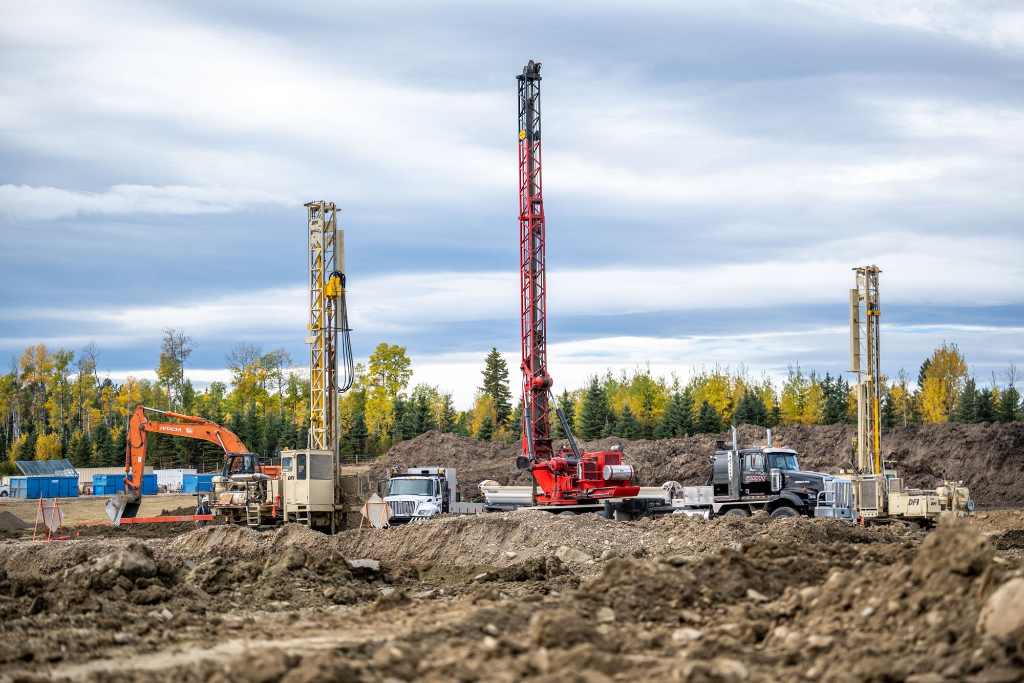 Construction site with drilling machinery, trucks, and equipment, surrounded by dirt and trees under a cloudy sky.