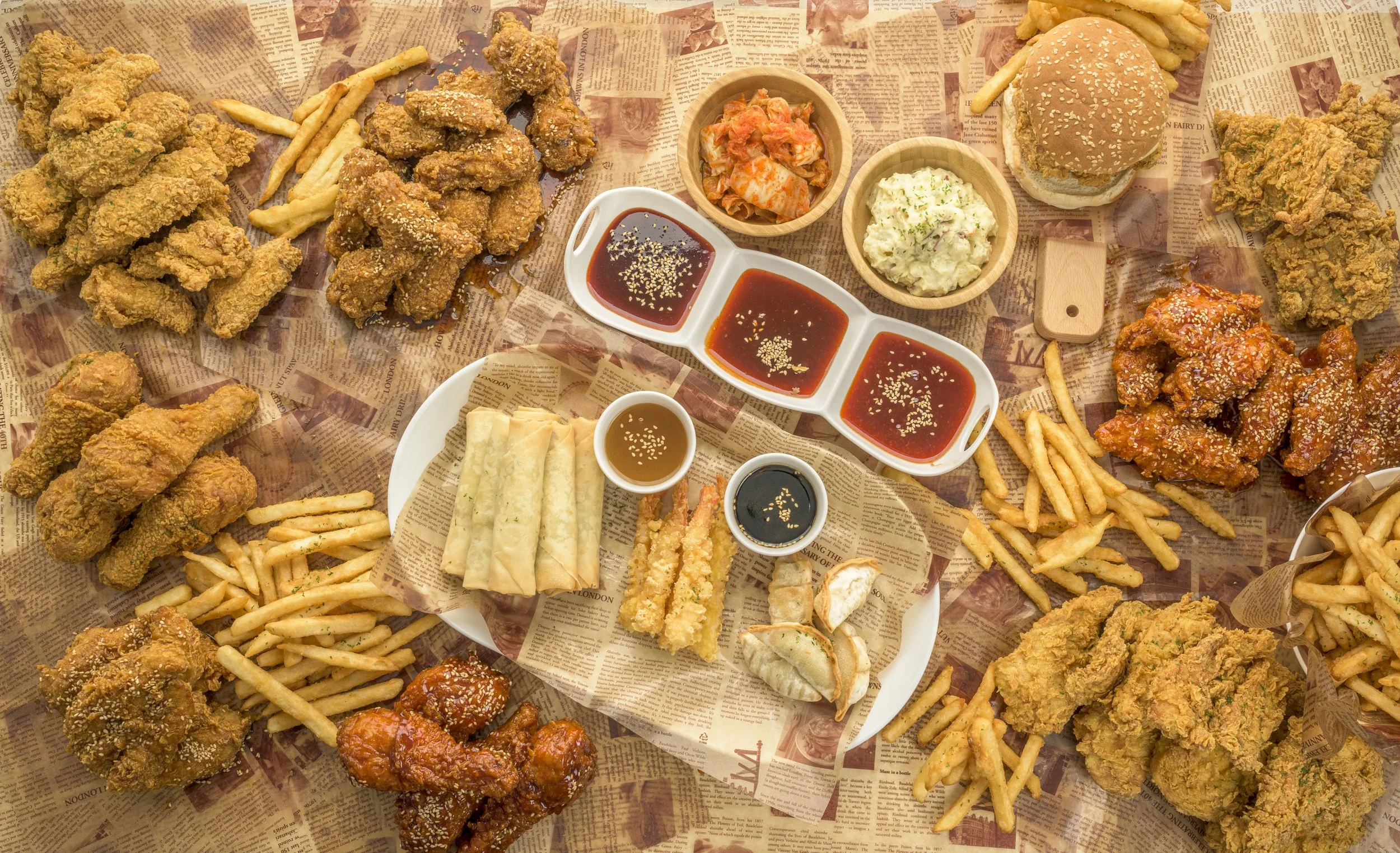 Assorted fried chicken, French fries, spring rolls, dumplings, and dipping sauces on newspaper-lined table.