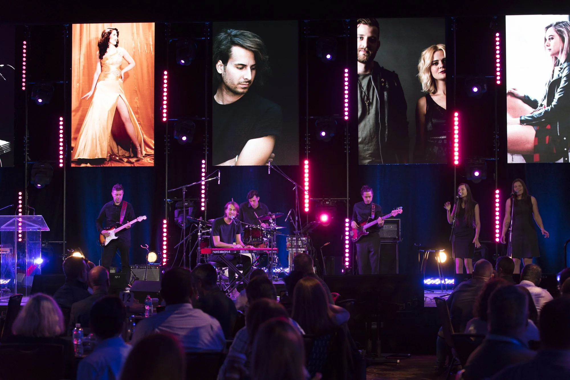 A musical performance on stage at a concert with band members playing guitar, keyboard, and drums, with four large screens above showing close-ups of individuals, and an audience watching the show.