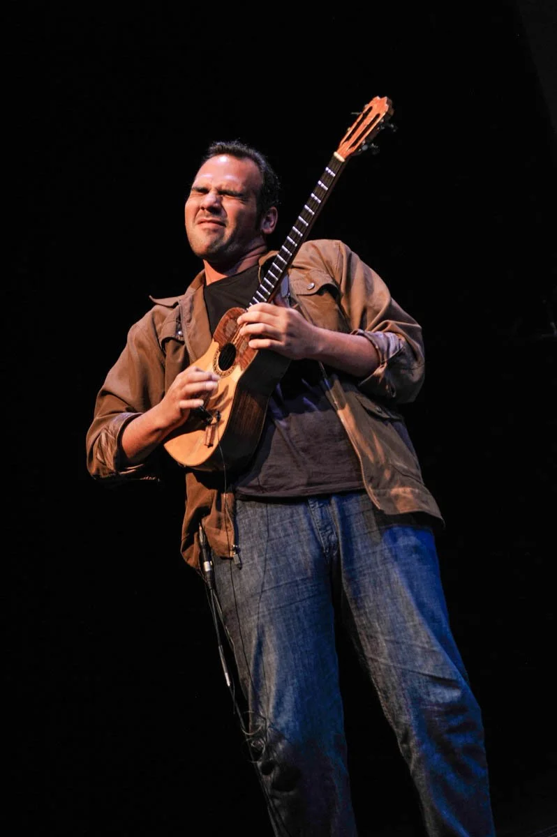 A man playing a small wooden guitar or ukulele on stage with a dark background.