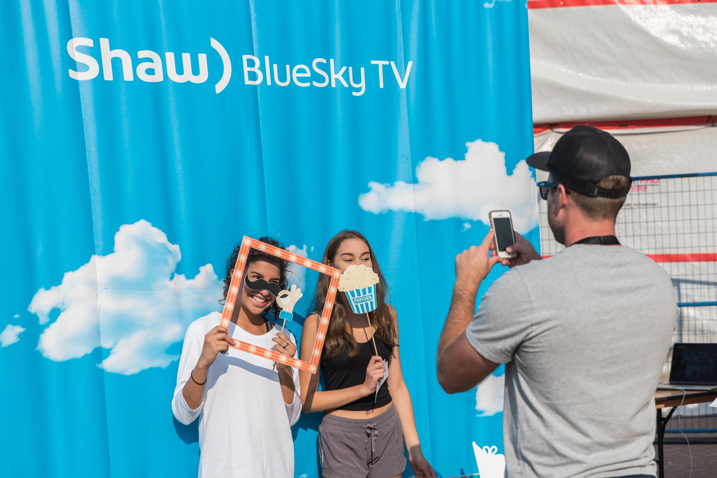 Two young women posing for a photo with fun props in front of a bright blue sky-themed backdrop with 'Shaw BlueSky TV' signage. One woman holds a mask with sunglasses and a mustache, while the other holds a tub of popcorn-shaped prop. A man in a gray