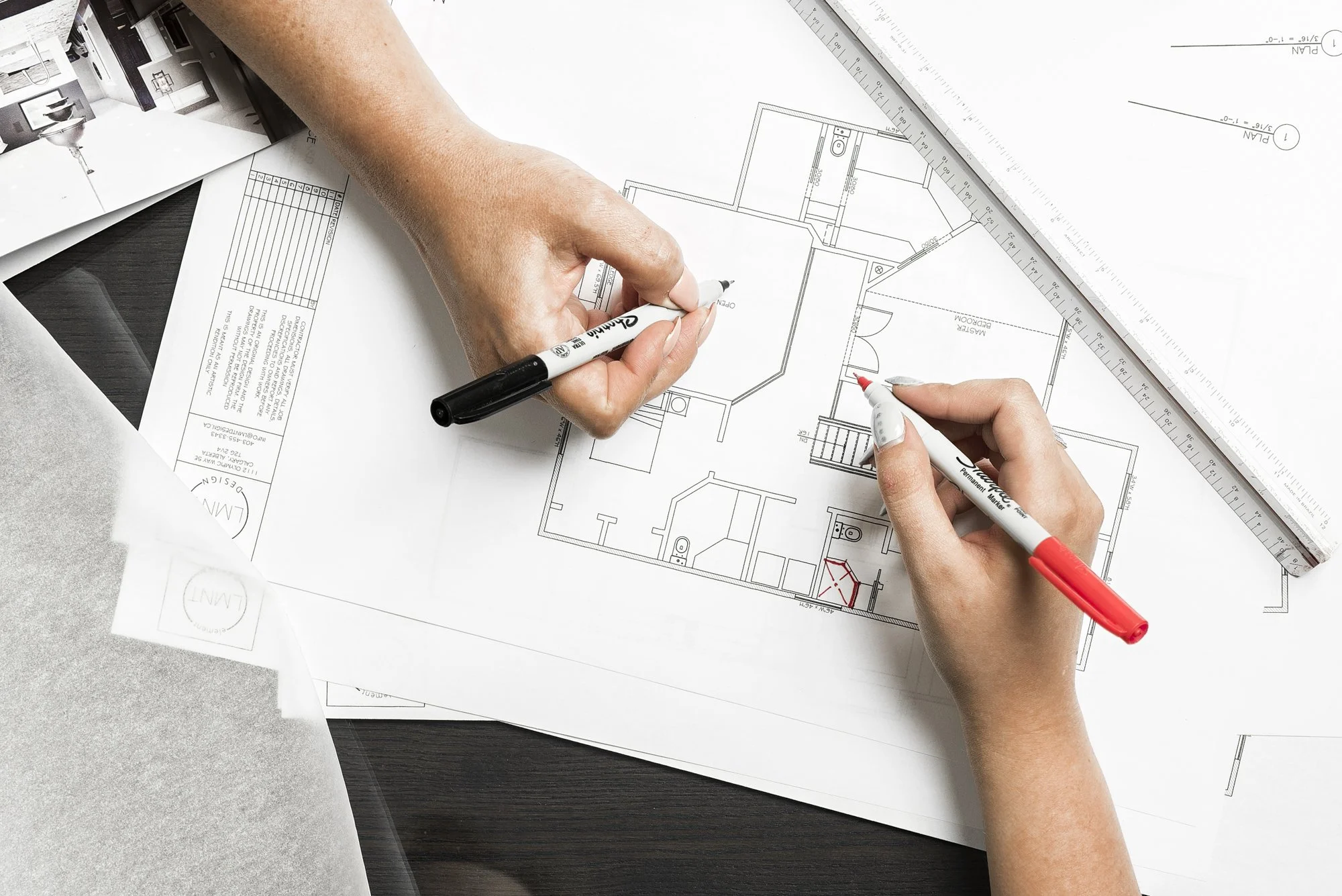 Two hands working on an architectural blueprint with black and red markers, on a dark wooden desk, with a large ruler and other documents.