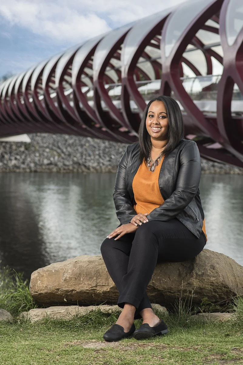 A woman sitting on a rock near water, in front of a modern, red, enclosed pedestrian bridge with unique design, smiling at the camera.