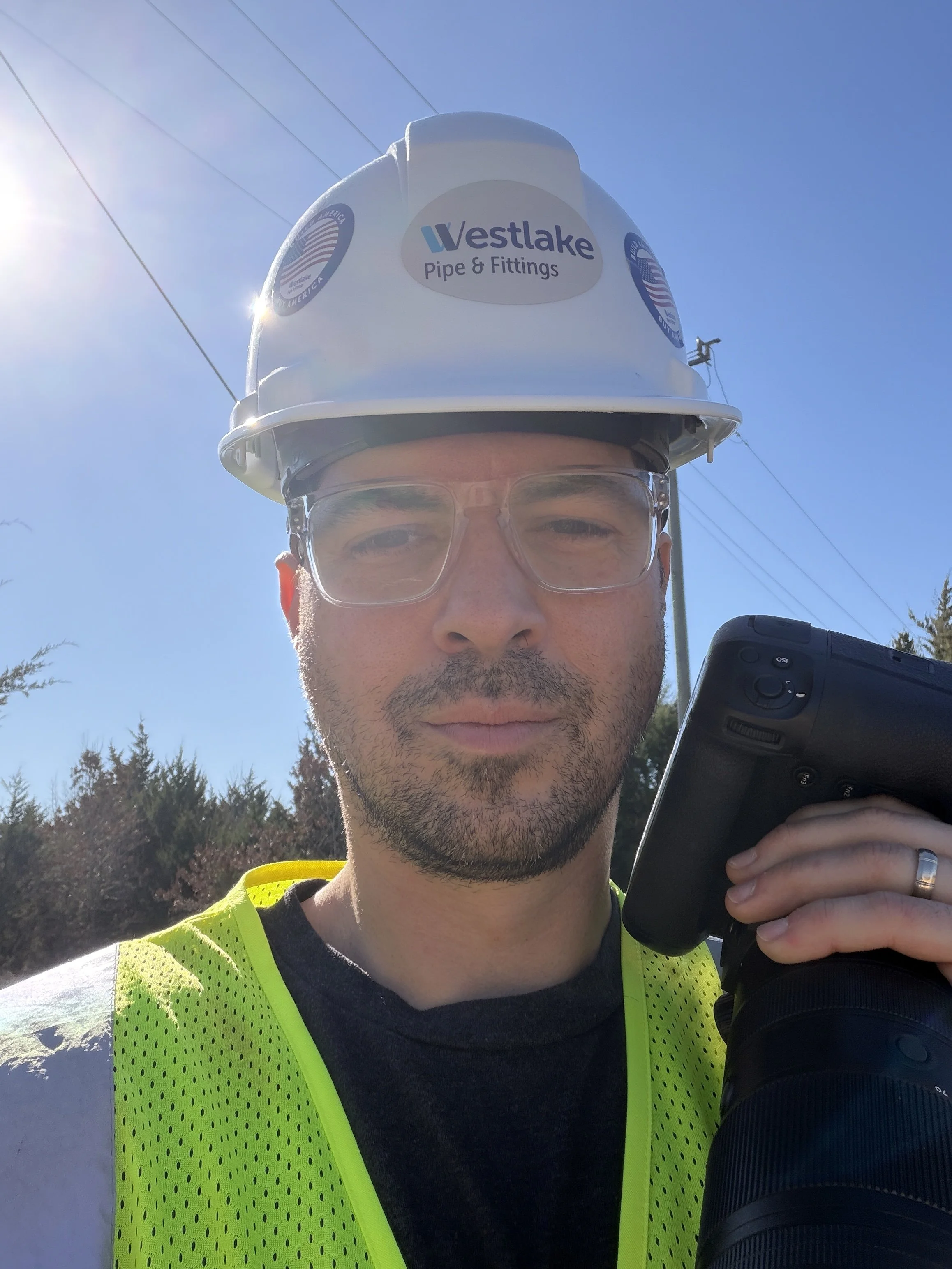 Construction worker wearing a white hard hat with 'Westlake Pipe & Fittings' logo, safety glasses, and a yellow safety vest, holding a camera or scanner in an outdoor setting with clear blue sky.