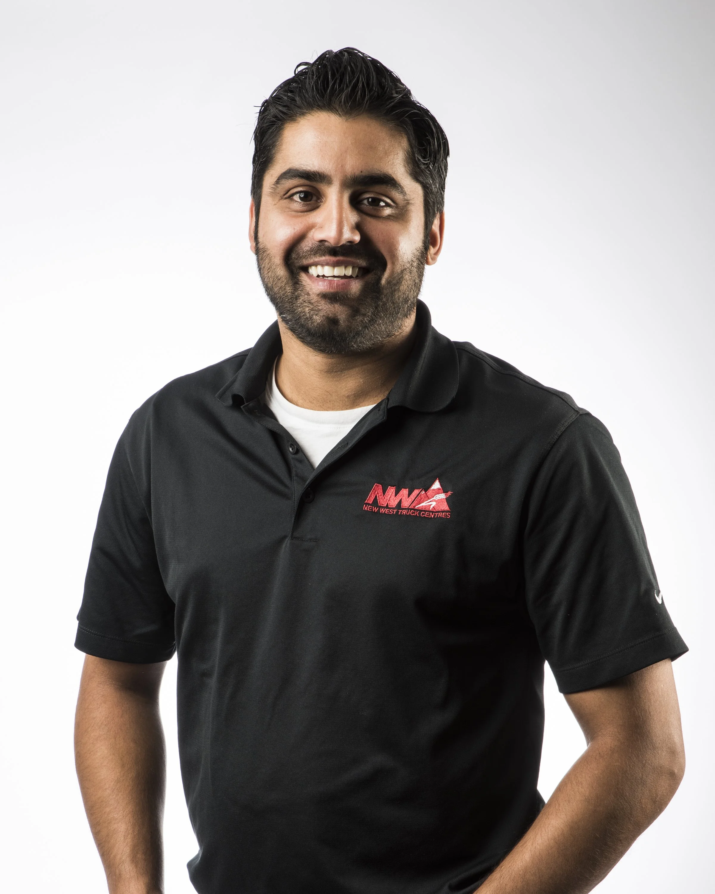A smiling man with dark hair and beard, wearing a black polo shirt with a red logo and white undershirt, standing against a white background.