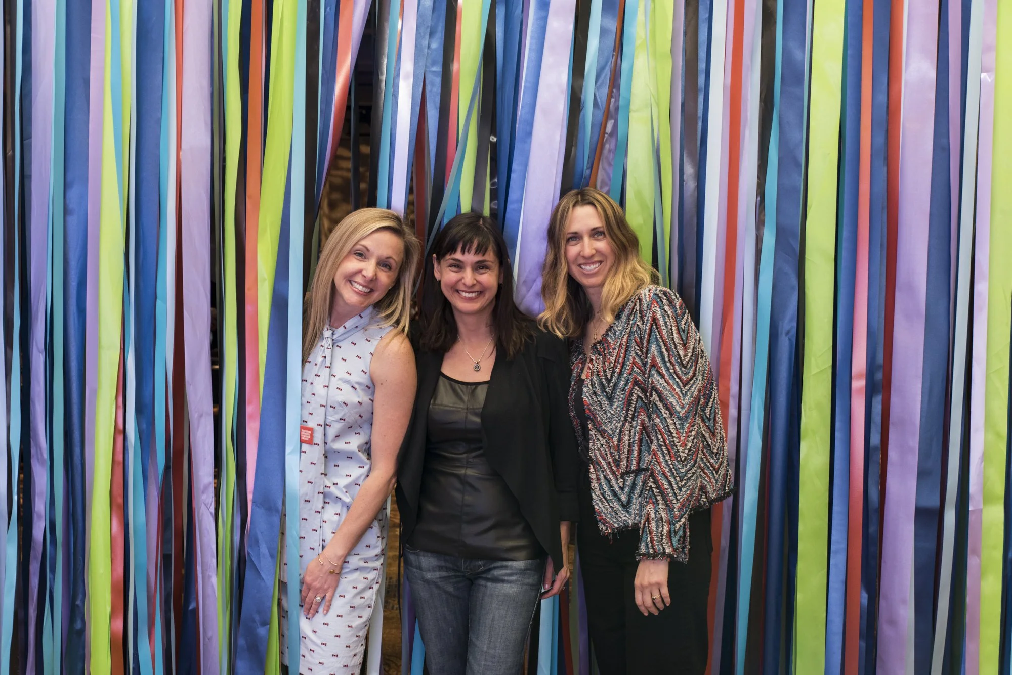 Three women smiling and standing close together in front of colorful streamers or ribbons.