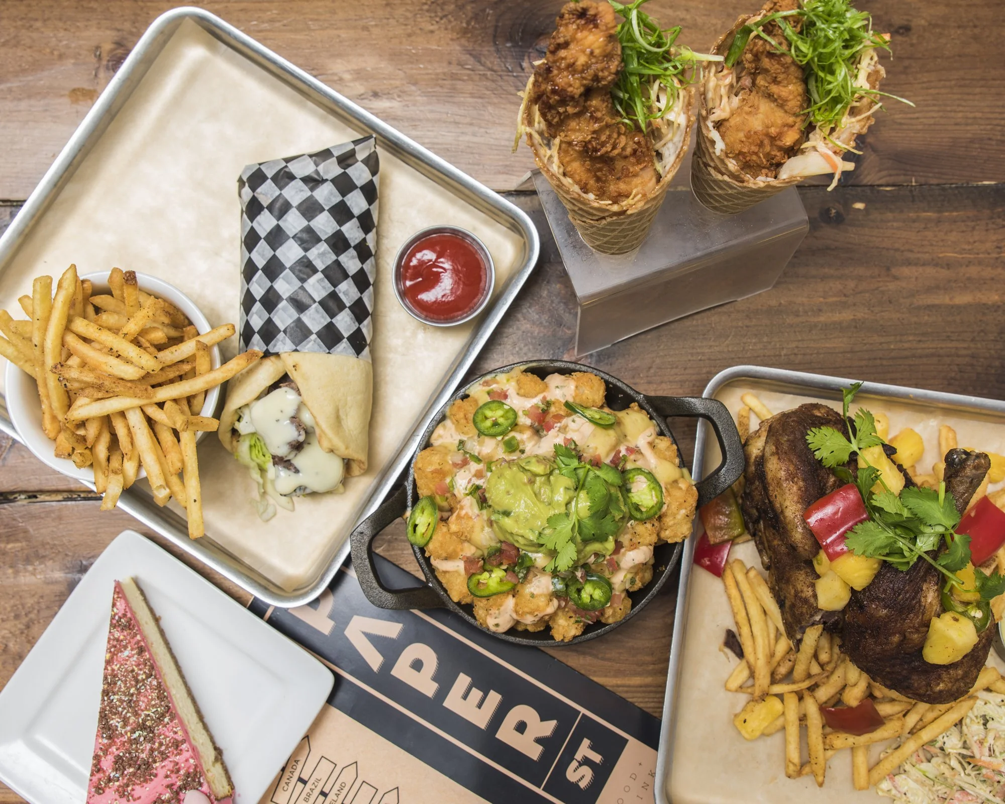 A variety of food on a wooden table, including French fries with a burger wrap, two ice cream cones with fried chicken and greens, a pot of fried cauliflower with jalapeños, a roasted chicken leg with vegetables and fries, and a slice of pink frosted