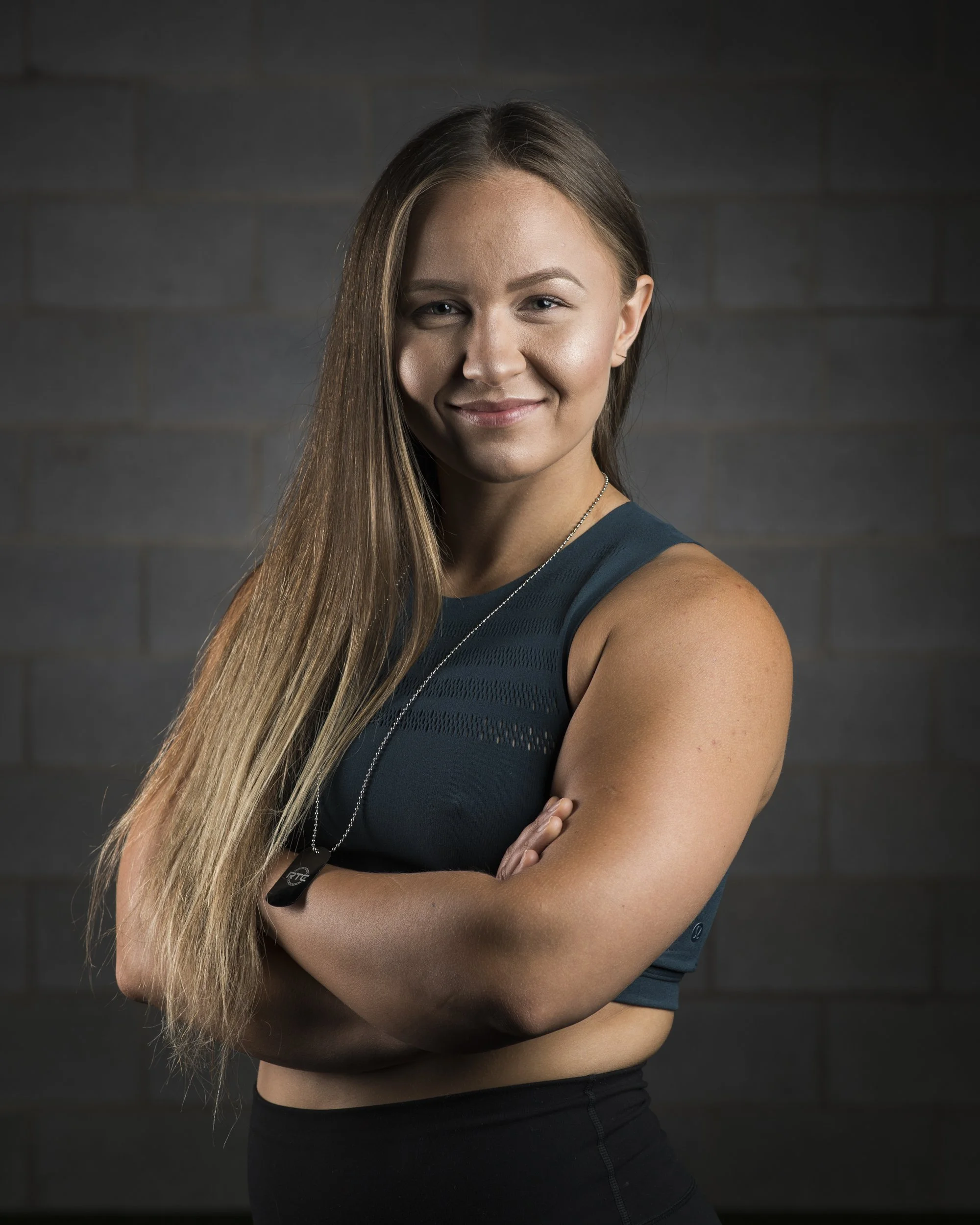 A fit young woman with long brown hair, smiling, standing in front of a dark brick wall, wearing a teal sleeveless athletic top and black workout leggings with her arms crossed, wearing a dog tag necklace.