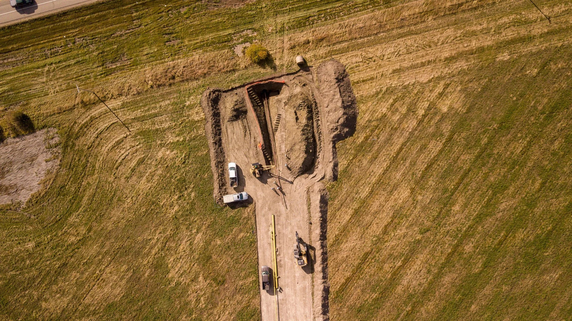 An aerial view of a construction site in a grassy field, showing workers, vehicles, and large machinery digging a rectangular hole.