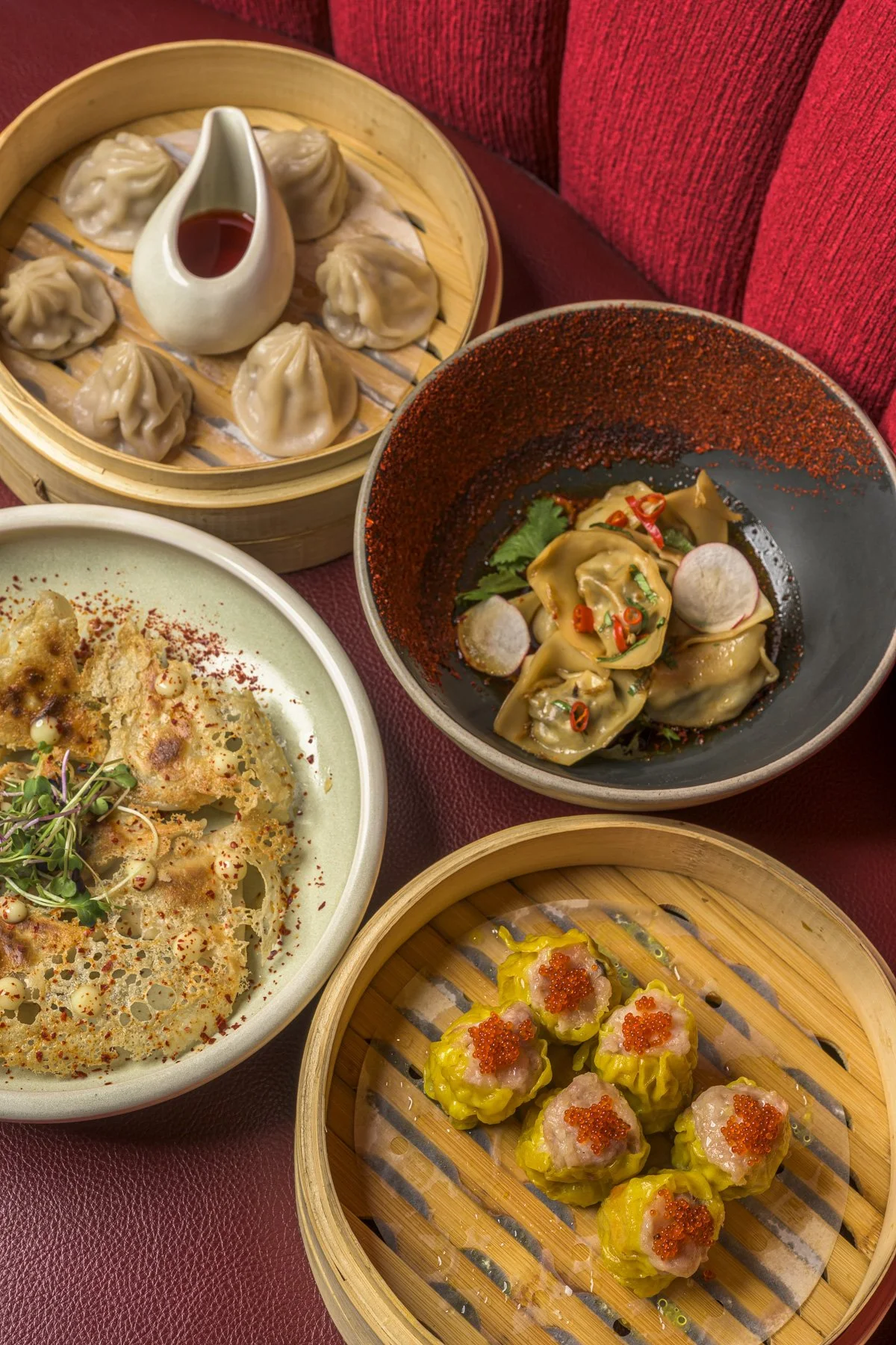 A variety of Asian dishes on a red table, including dumplings with red sauce in a bamboo steamer, dumplings in a black bowl garnished with herbs and chilies, crispy fried dumplings topped with sprouts on a green plate, and siu mai dumplings with fish