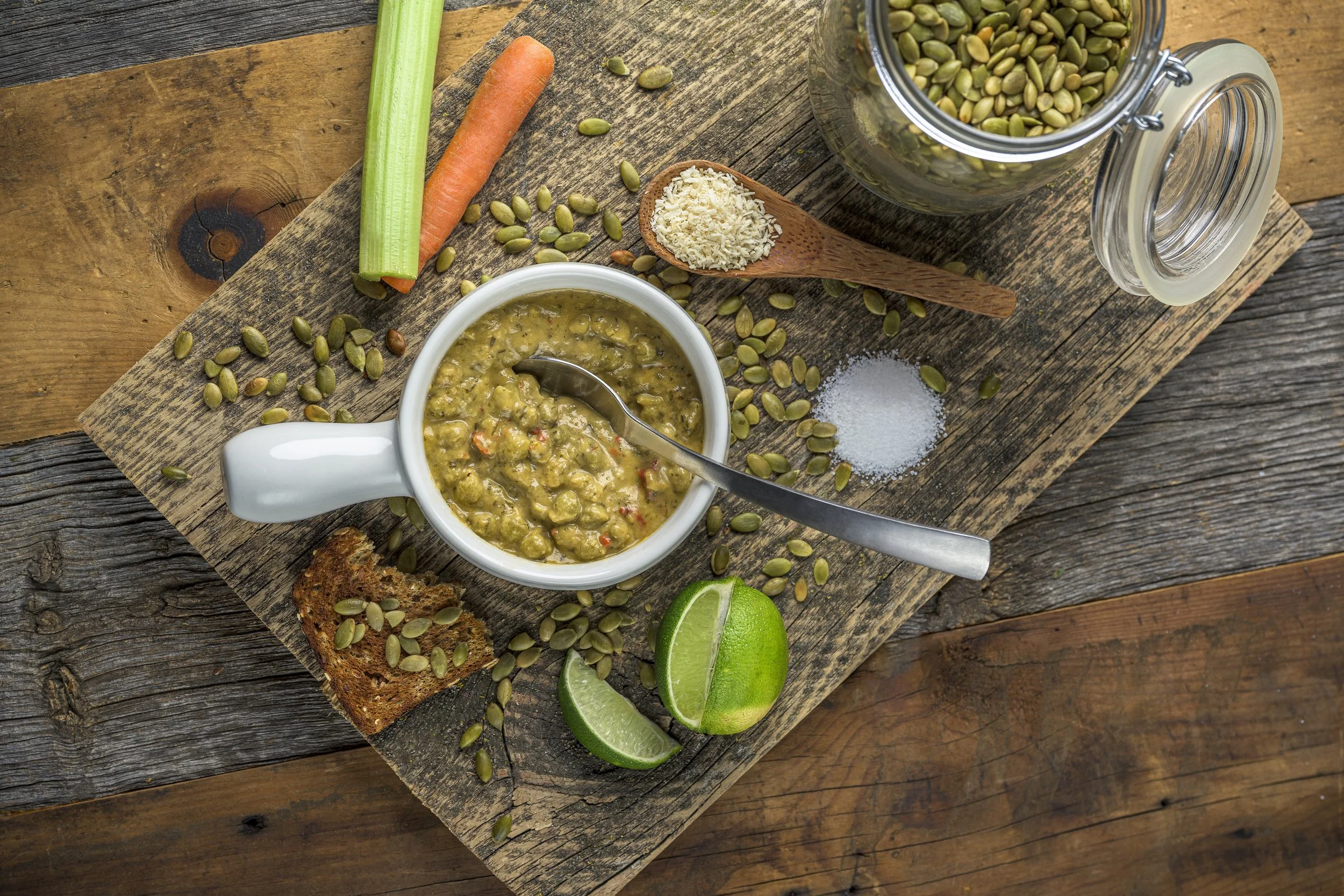 A rustic wooden table holds a variety of foods and ingredients, including a mug of guacamole with a spoon, lime wedges, a piece of toasted bread with seeds, a carrot, a celery stick, a jar of pumpkin seeds, a spoonful of grated cheese, and scattered 