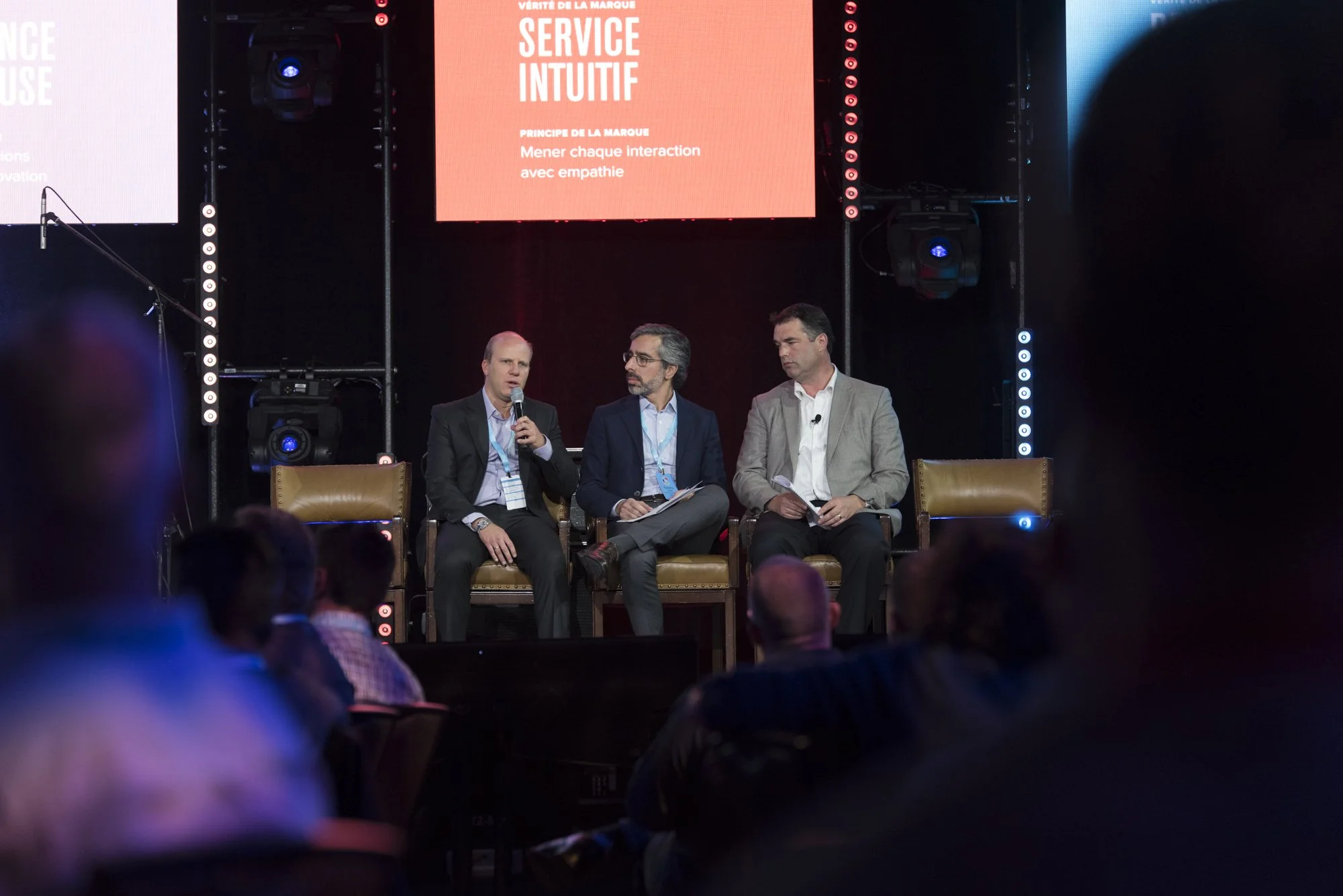 Three men sitting on stage in a panel discussion at a conference, with a large screen behind them displaying French text about intuitive service and empathy.