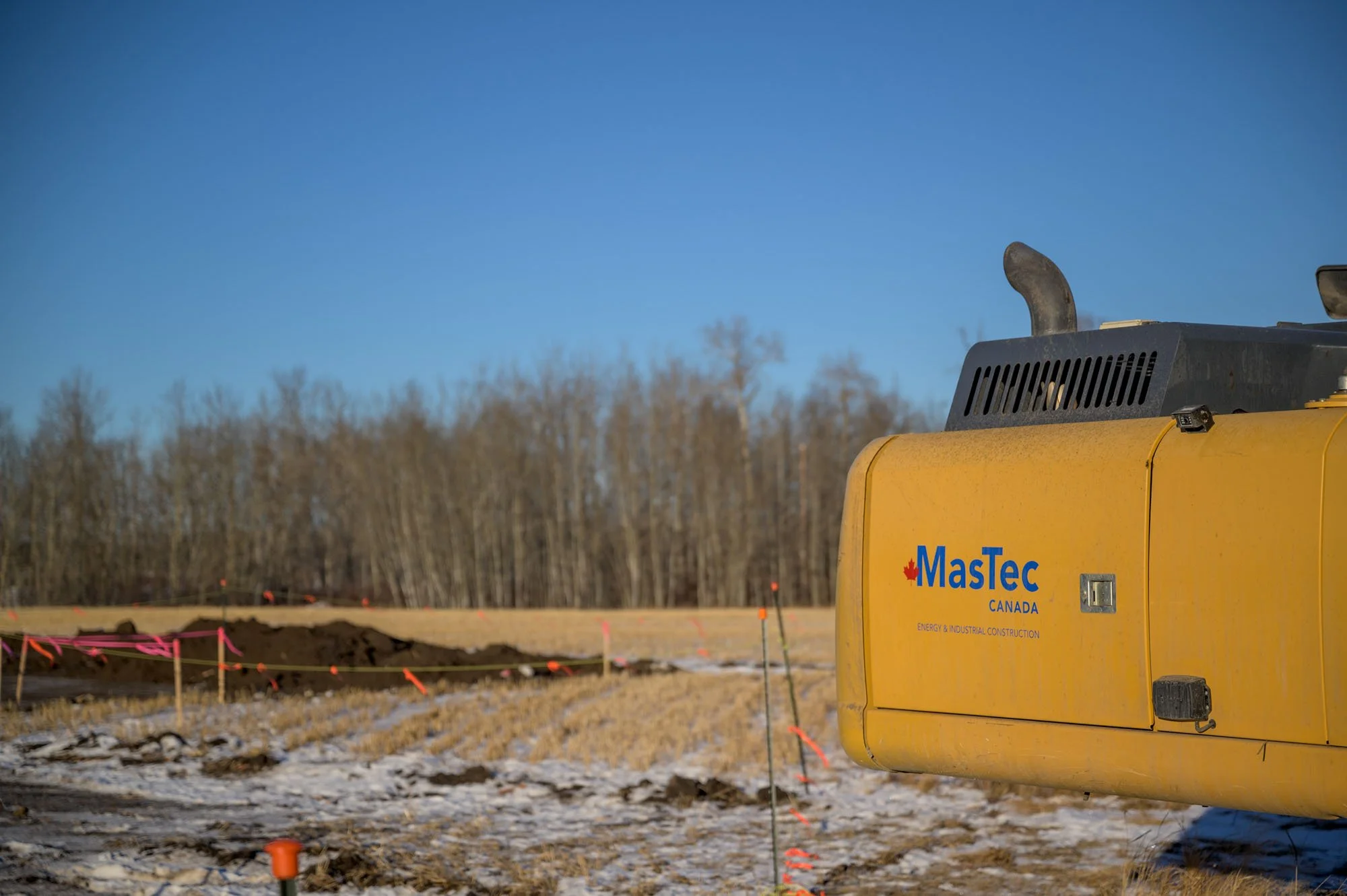 Close-up of a yellow construction vehicle with the words 'MaStEc CANADA' and 'ENERGY & INDUSTRIAL CONSTRUCTION' on it, at a construction site with dirt and snow, pink and orange markers, and a background of leafless trees and a clear blue sky.
