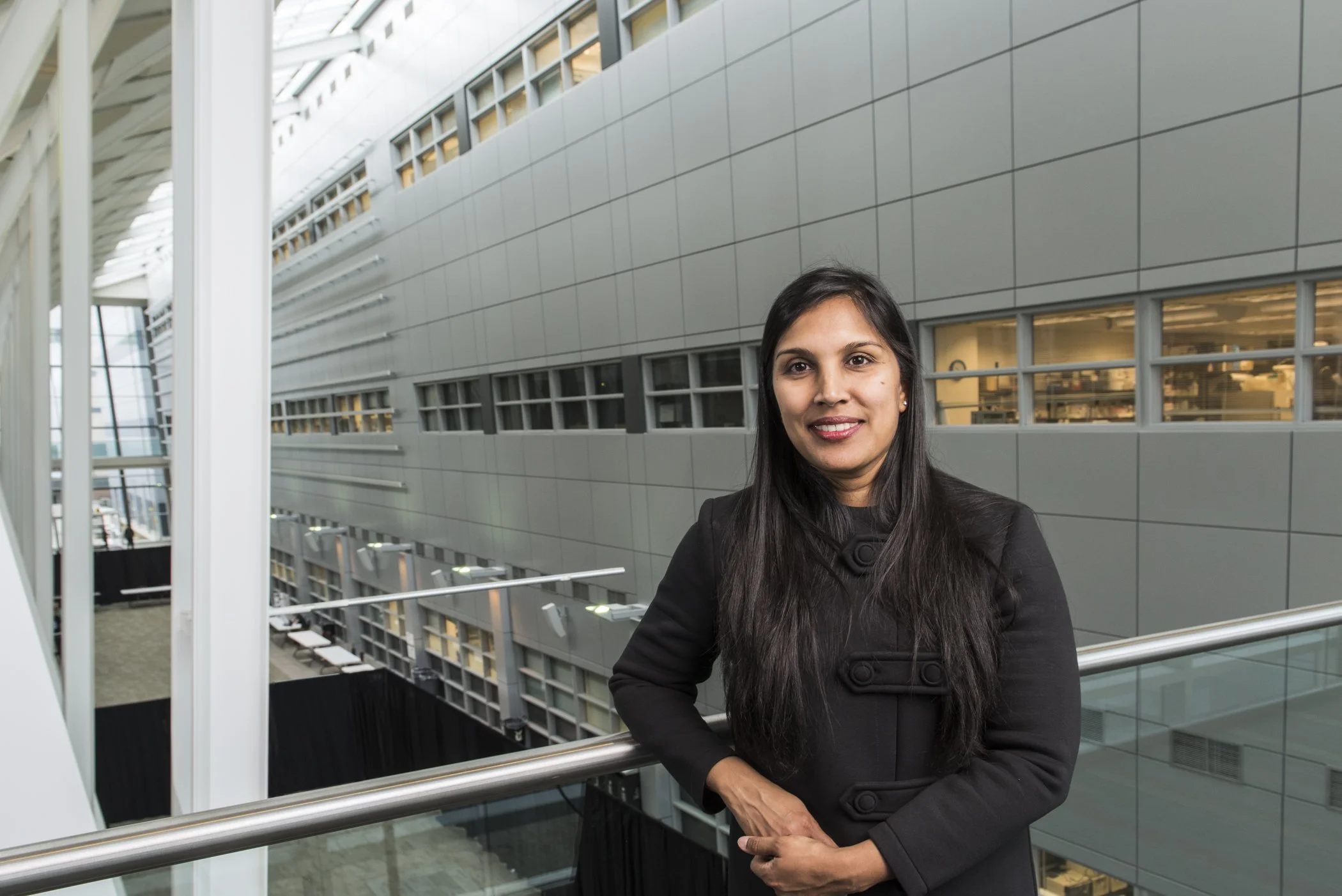 A woman with long black hair wearing a black coat, standing indoors in front of a large modern building with a glass and metal structure.