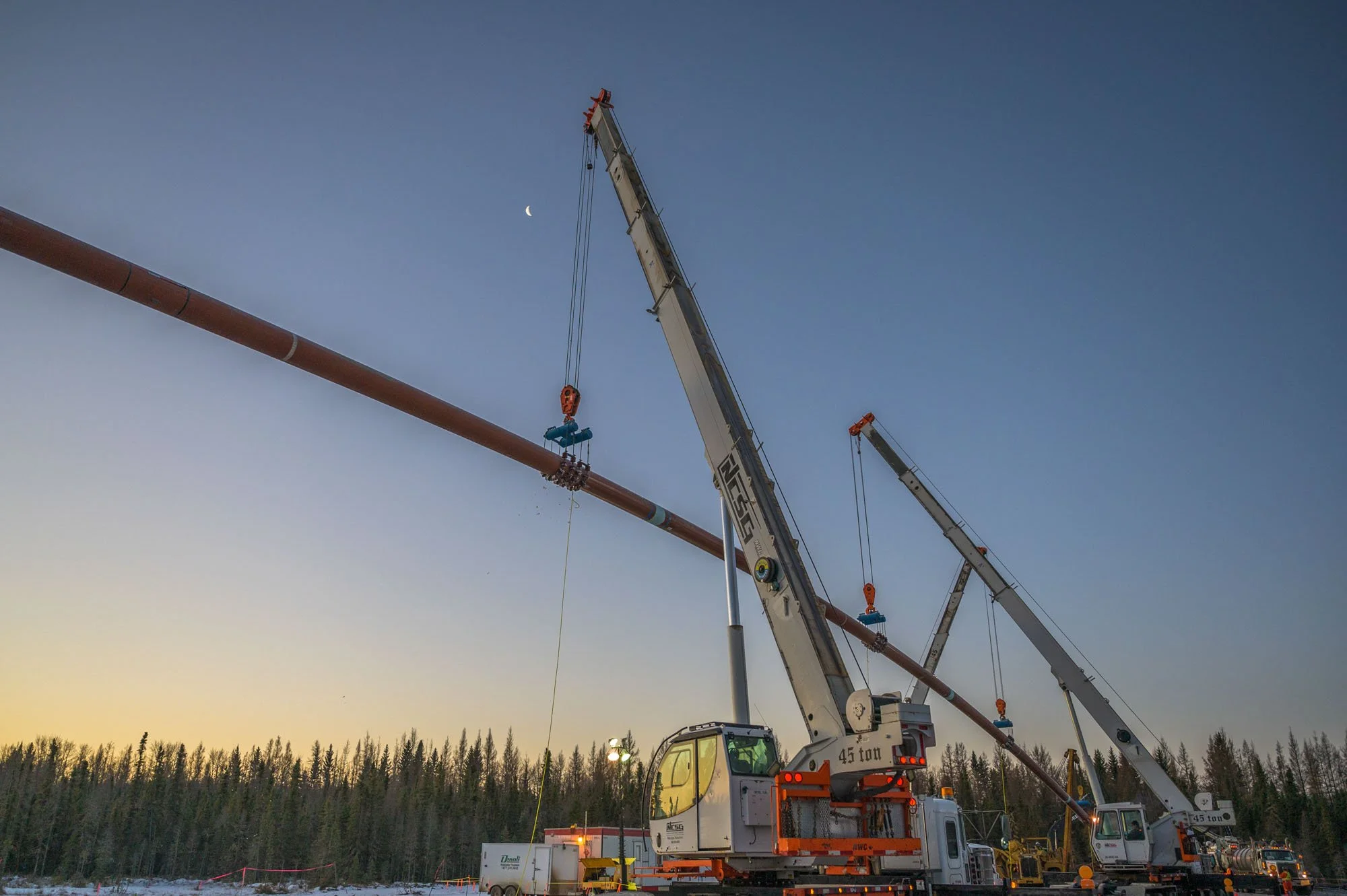 Two cranes lifting a large pipeline against a sunset sky with a crescent moon and trees in the background.