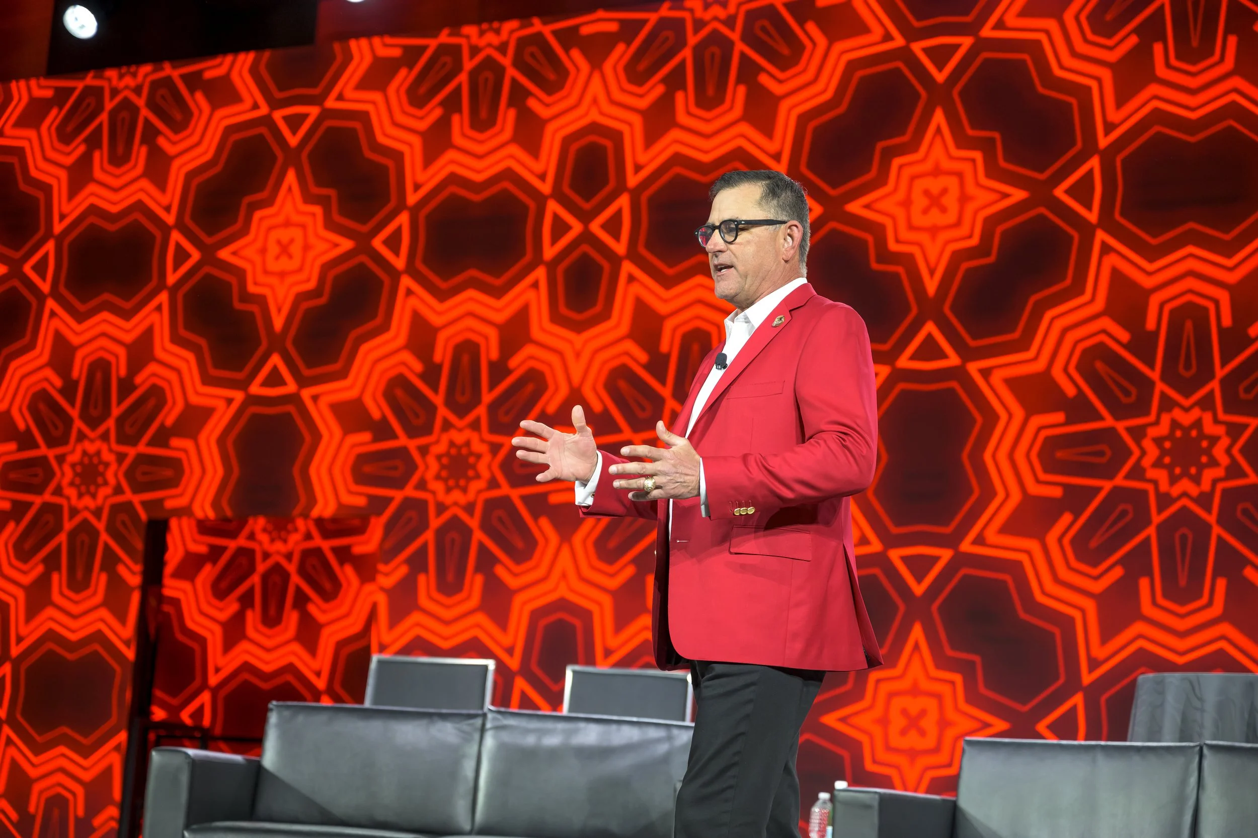 A man wearing a red blazer and glasses is standing on stage, speaking with hand gestures, in front of an intricate red and black geometric patterned background.