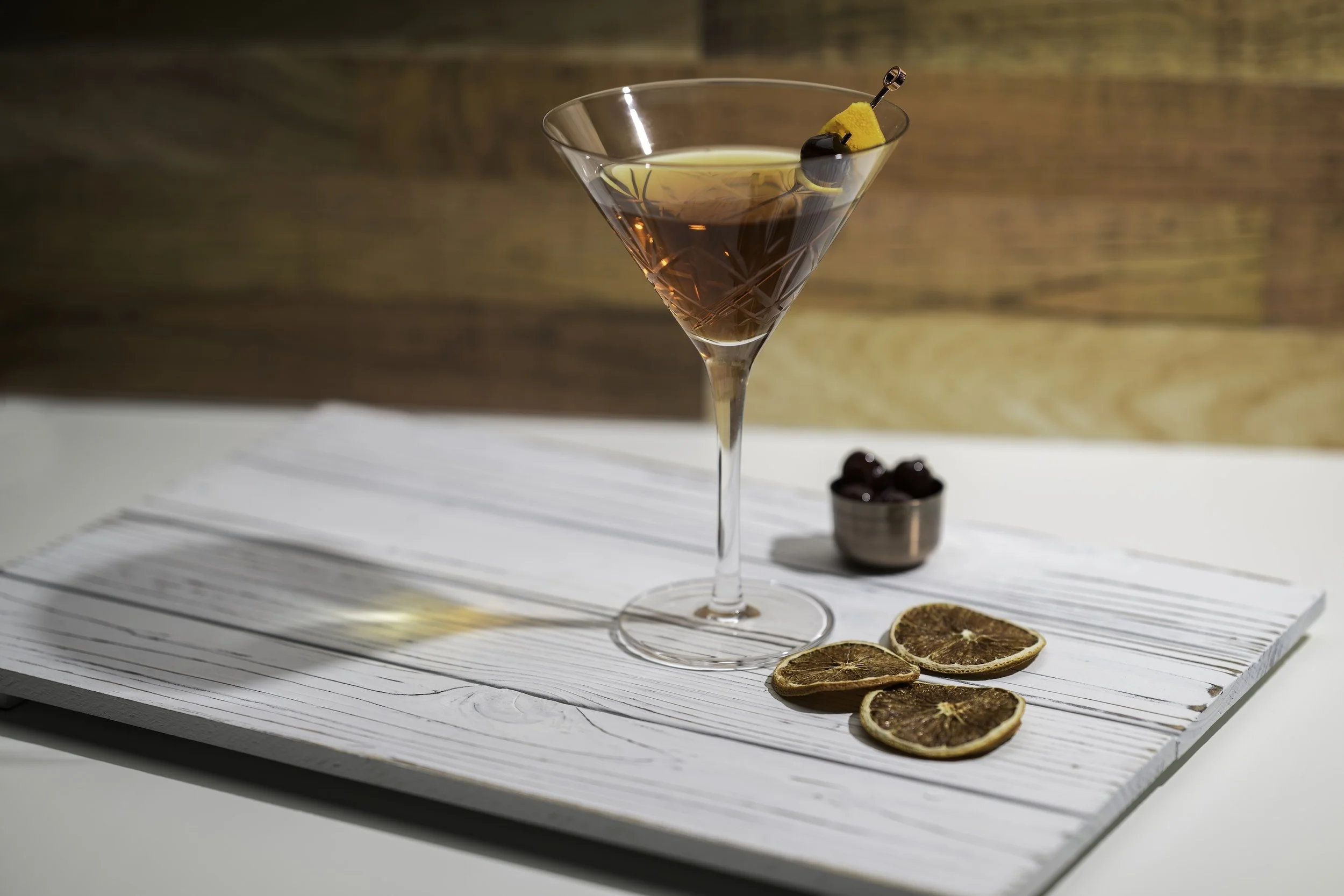 An elegant cocktail in a martini glass garnished with a cherry and orange peel, placed on a white wooden surface with dried orange slices and a small bowl of cherries nearby.