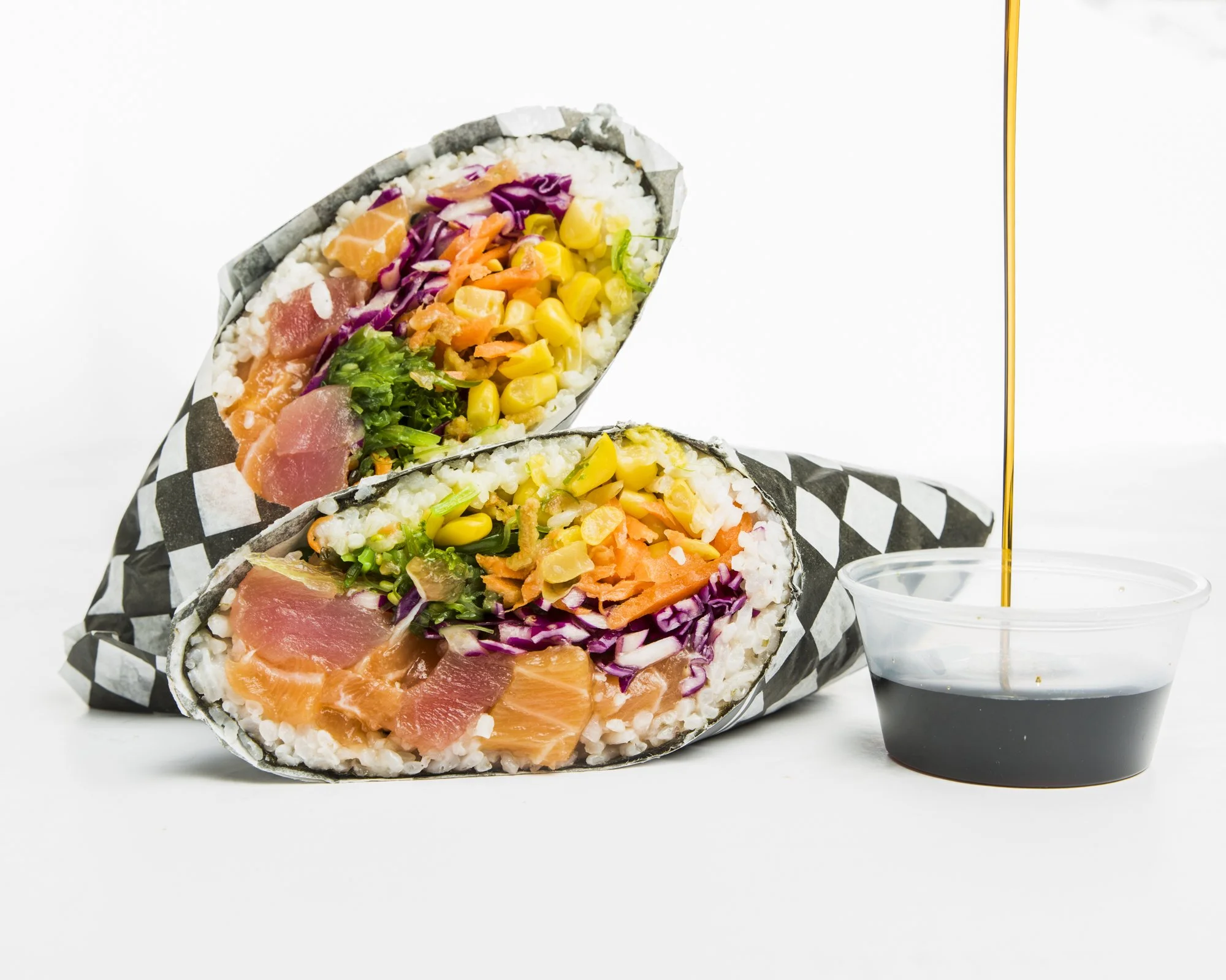 Two sushi burritos with colorful vegetables and raw fish, with soy sauce being poured over them.