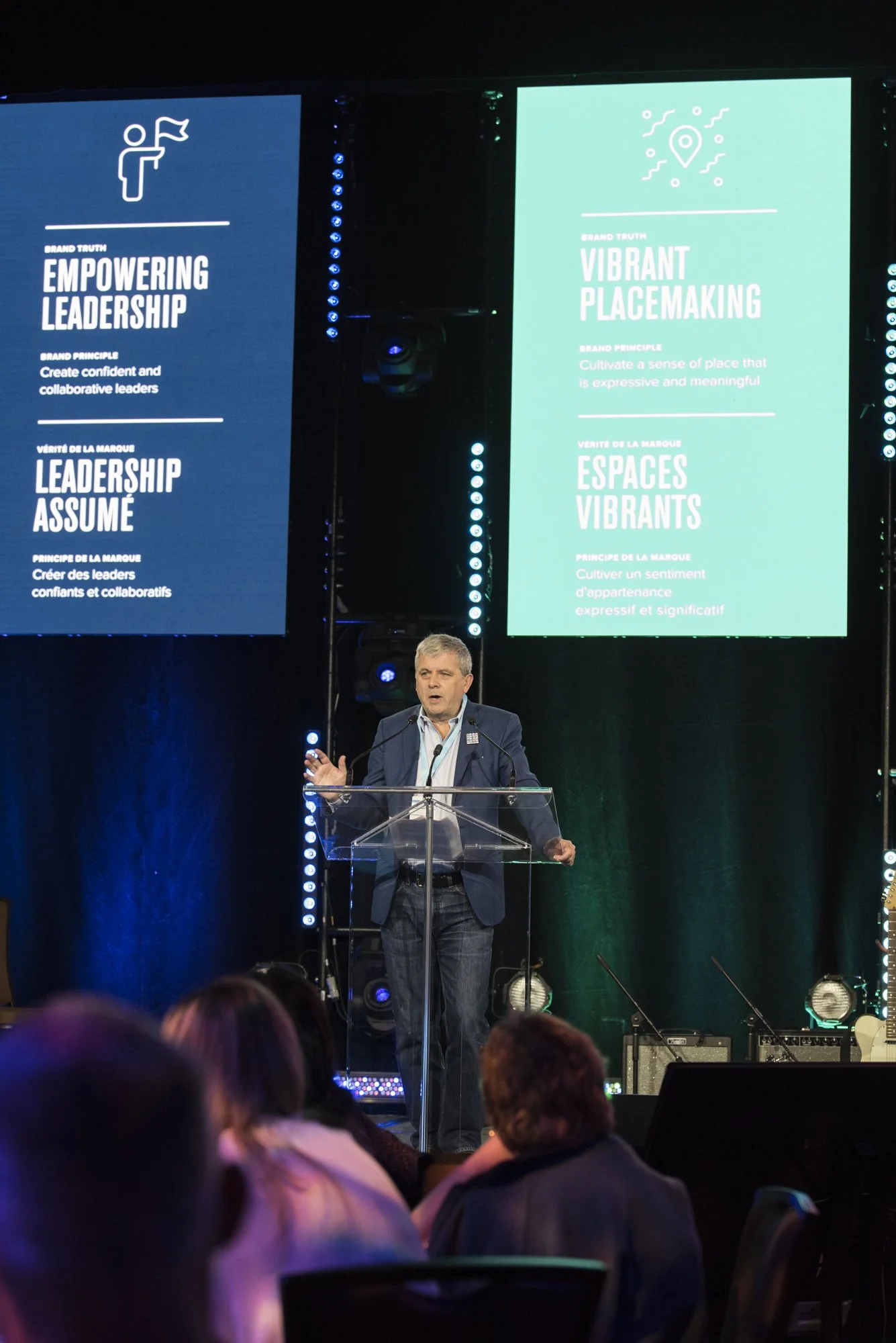 A man in a suit speaking at a podium during a conference with large screens behind him displaying branding principles in English and French, including 'Empowering Leadership' and 'Vibrant Placemaking'.