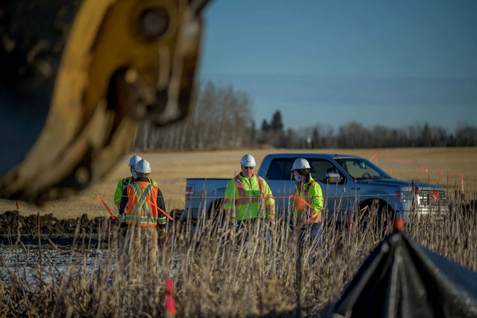 Construction workers in high-visibility safety vests and white helmets working in a field, with a pickup truck in the background. The photo is taken from beneath a piece of heavy machinery.