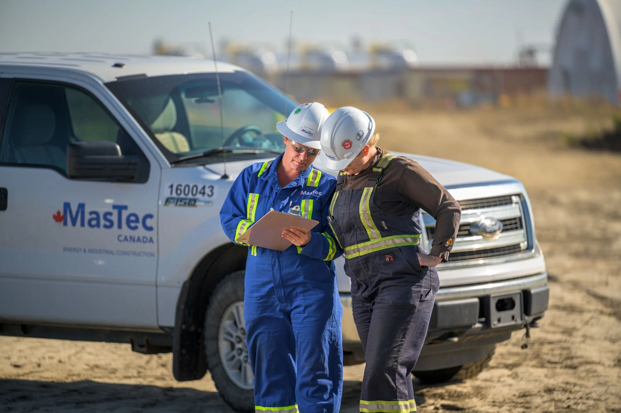 Two energy workers in safety gear discussing in front of a MasTec service vehicle at an outdoor work site.
