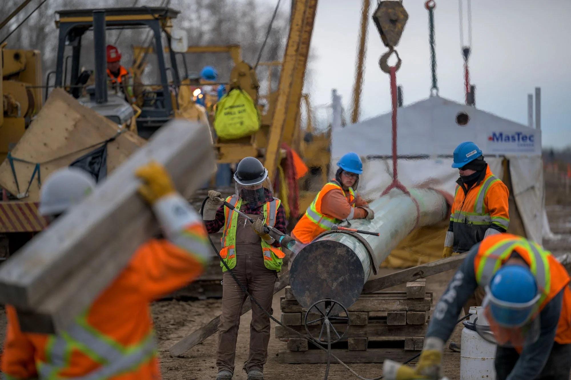 Construction workers in safety vests and helmets installing a large pipe at a construction site with heavy machinery in the background.
