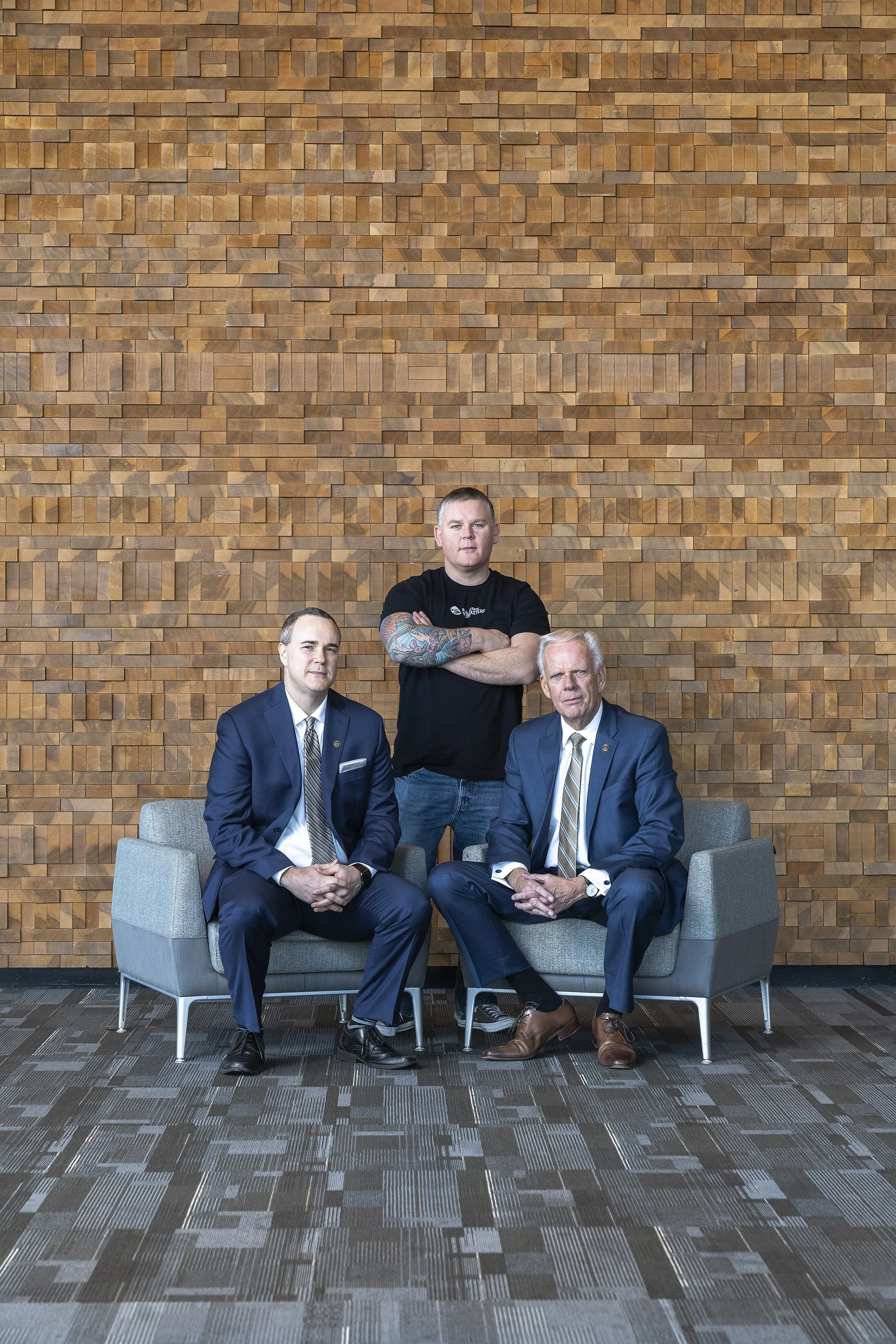 Three men, two in suits and one in casual wear, pose for a photo in front of a uniquely textured wooden wall. The men in suits are seated on gray chairs, while the casually dressed man stands behind them with his arms crossed.