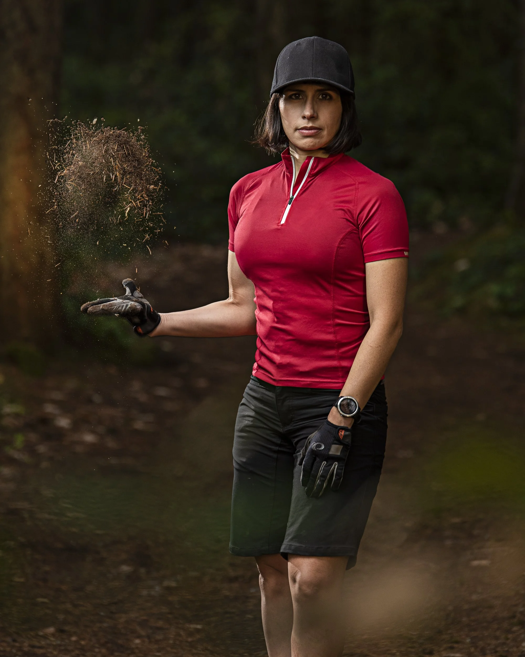 A woman in athletic gear stands in a wooded area, holding dirt and debris in one hand, with a confident expression.