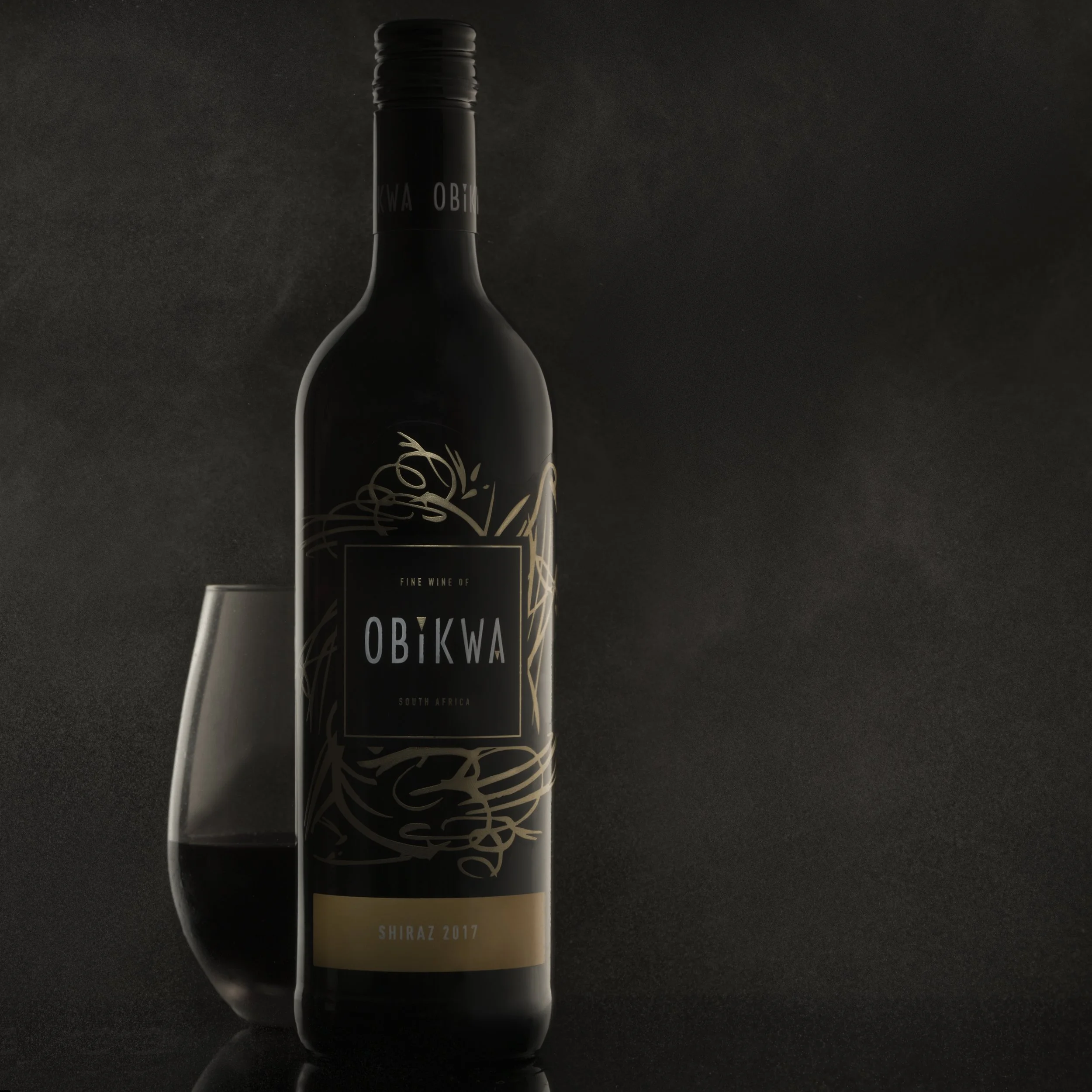 A dark wine bottle labeled 'Obikwa South Africa Shiraz 2017' with a transparent wine glass filled with red wine beside it, set against a dark background.